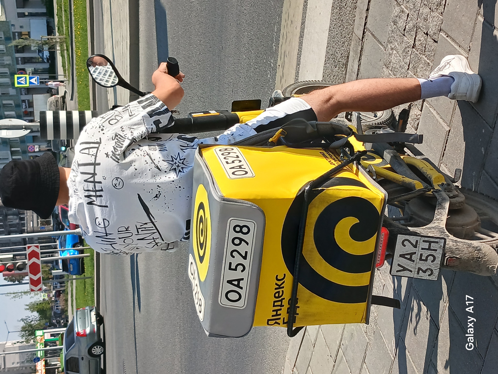 A man is riding a bicycle on a city street. He is wearing a white shirt with black print, black shorts with white accents, white socks, and white sneakers. He is also wearing a black bucket hat. The bicycle has a large yellow delivery box on the back with the Yandex logo and the number OA5298. The license plate on the bicycle reads YA235H. The background shows a city street in Yekaterinburg, Russia, with buildings, trees, traffic lights, and other vehicles.