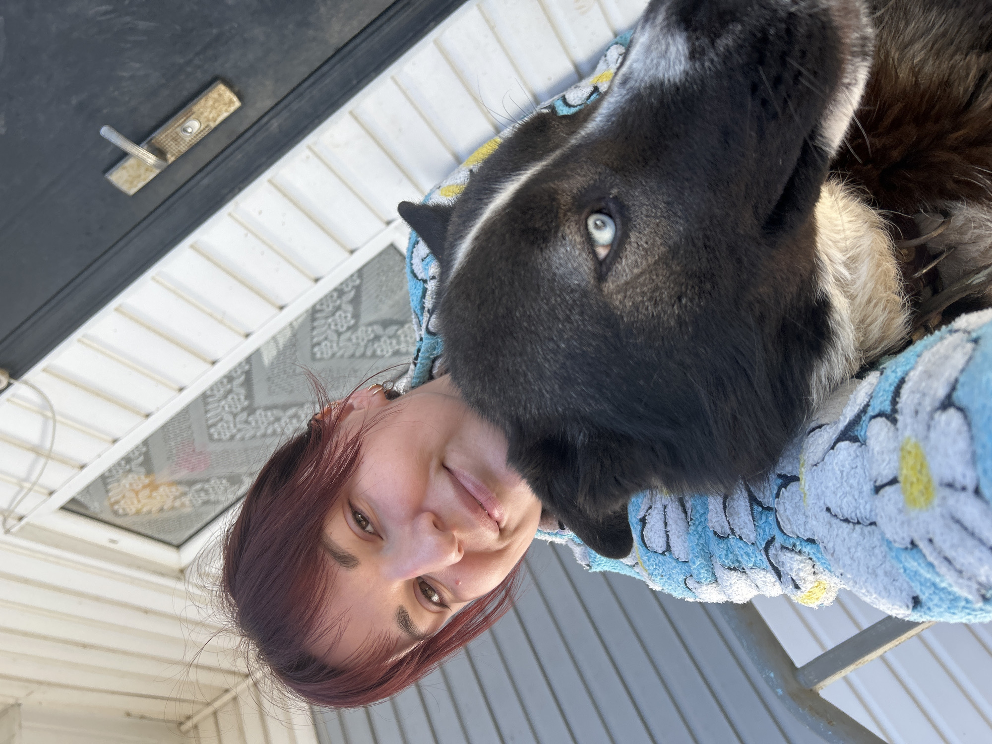 A woman with auburn hair is holding a black and white husky-type dog in her arms outdoors, on what appears to be a porch or deck. The dog's head is close to the woman's face, and its eye is light blue. The woman is wearing a blue and white floral pattern fabric. Behind them is a white-sided building with a dark door and a security screen on the window. The location is Фоки, Russia.