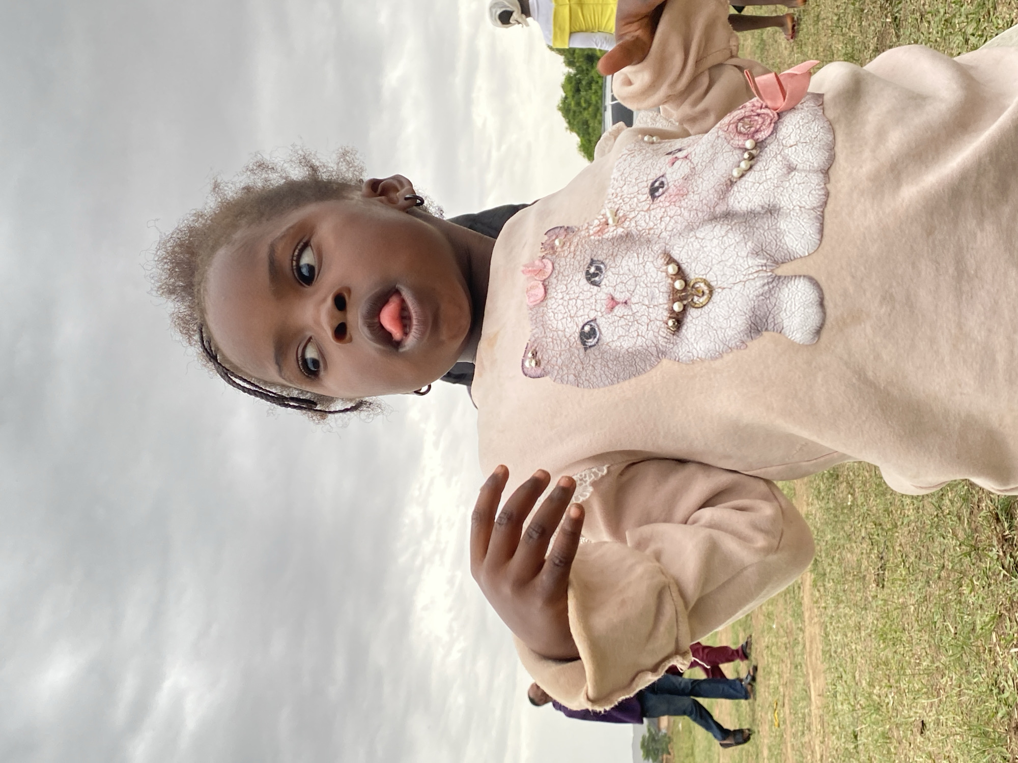 A young, dark-skinned child with braided hair, dressed in a light brown sweatshirt with a cat image, is sticking out her tongue and looking at the camera. She is outdoors on grassy terrain with other people visible in the background. The sky is overcast with gray clouds. The location is Kuru Dungus, Nigeria.