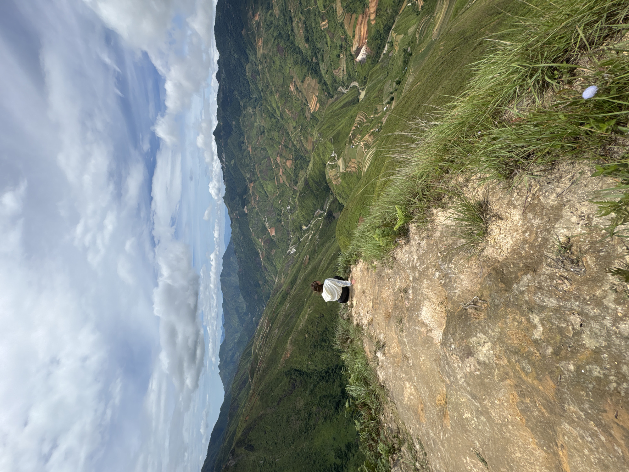 The image captures a person from behind, seated on a rocky outcrop at the edge of a steep, verdant mountain landscape. The person is wearing a white hooded jacket and dark pants, gazing out at the expansive vista of rolling green hills and valleys. The terrain is characterized by terraced fields, winding roads, and patches of exposed earth, indicative of agricultural activity and human presence in the mountainous region. The sky above is partly cloudy, with dramatic formations of white and grey clouds against a blue backdrop. The overall scene suggests a remote and natural setting, likely in the highlands of Ta Xua, Vietnam, a location known for its breathtaking mountain scenery and tea plantations. The foreground is dominated by the rough texture of the rock and the lush green grass growing along the cliff edge.