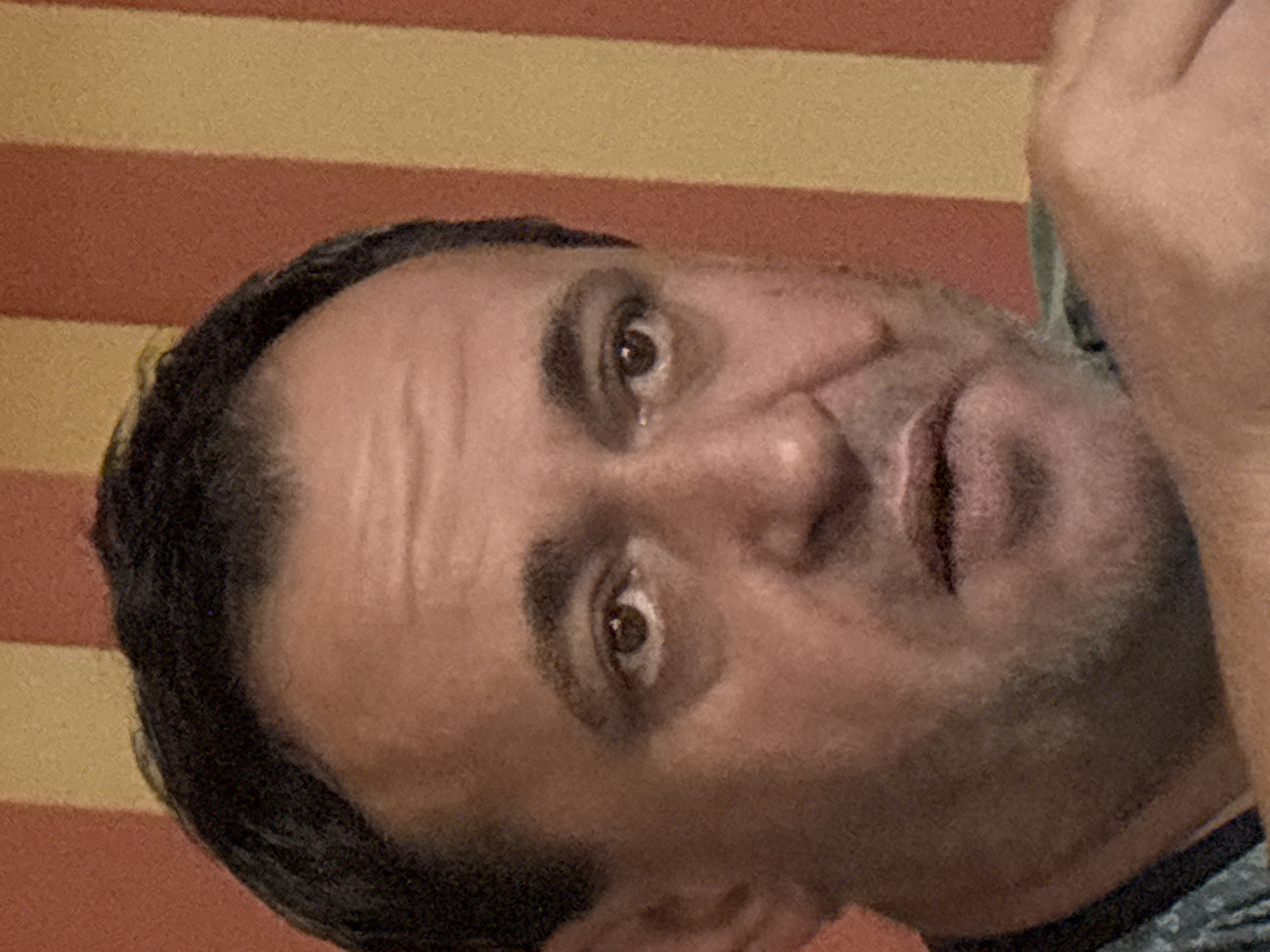 A close-up, waist-up, head-and-shoulders studio portrait depicts a middle-aged Caucasian man with dark hair, light brown eyes, and light skin. He has a shaved head with some dark stubble on his face, a faint five o'clock shadow, and prominent eyebrows. His forehead shows visible wrinkles, and his eyes are wide open, looking directly at the camera. The background consists of vertical stripes in alternating shades of red and yellow. The man is wearing a dark t-shirt with a subtle pattern and a light green collar. His left hand is visible resting on his shoulder. The image was taken in Tervel, Bulgaria.
