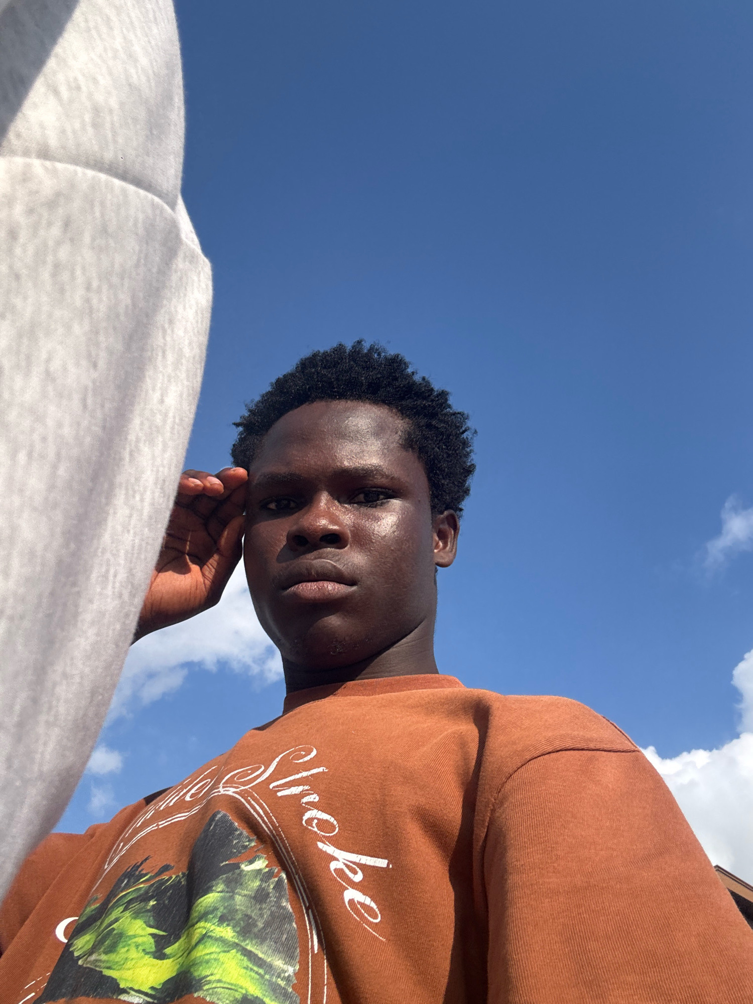 In Ihiagwa, Nigeria, a young Black man is captured in a low-angle selfie against a bright blue sky with scattered clouds. He has dark, curly hair and is looking directly at the camera. His right hand is raised to his forehead, obscuring part of his face. He is wearing a brown crewneck sweatshirt with white text that appears to read "The Smoke" and a graphic design on the front. To the left of the frame, a large, soft, off-white object, possibly a piece of clothing or fabric, takes up a significant portion of the image, its texture suggesting a thick material. The lighting indicates it is daytime, with sunlight illuminating the man's face and casting a slight sheen on his skin.