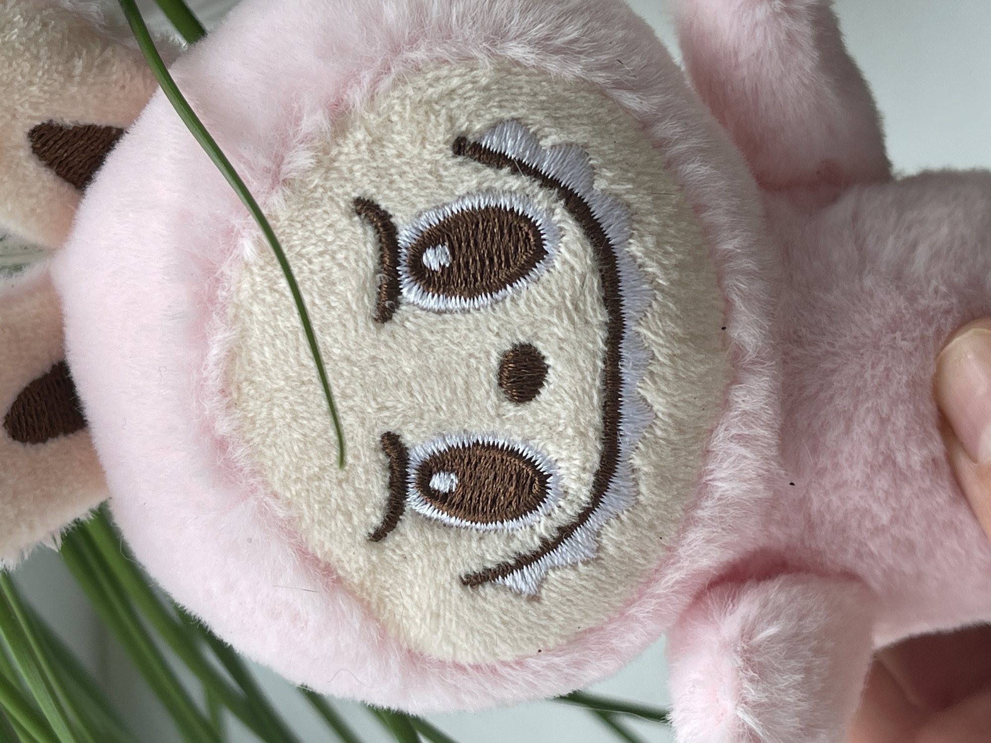 A close-up, eye-level shot shows a pink plush toy rabbit. The toy is positioned in the foreground, with its face filling most of the frame. It has large, embroidered brown eyes with white highlights, curved brown eyebrows, and a wide, stitched smile with white teeth. The toy's head is covered in a pink hood, and its ears, also pink, have brown embroidered inner sections. A thin green blade of grass runs diagonally across the toy's face, from the upper left to the lower right. In the background, out of focus, are more green blades of grass and a light gray surface. A human finger is visible at the bottom left of the frame, holding the toy. The location is not discernible from the image.