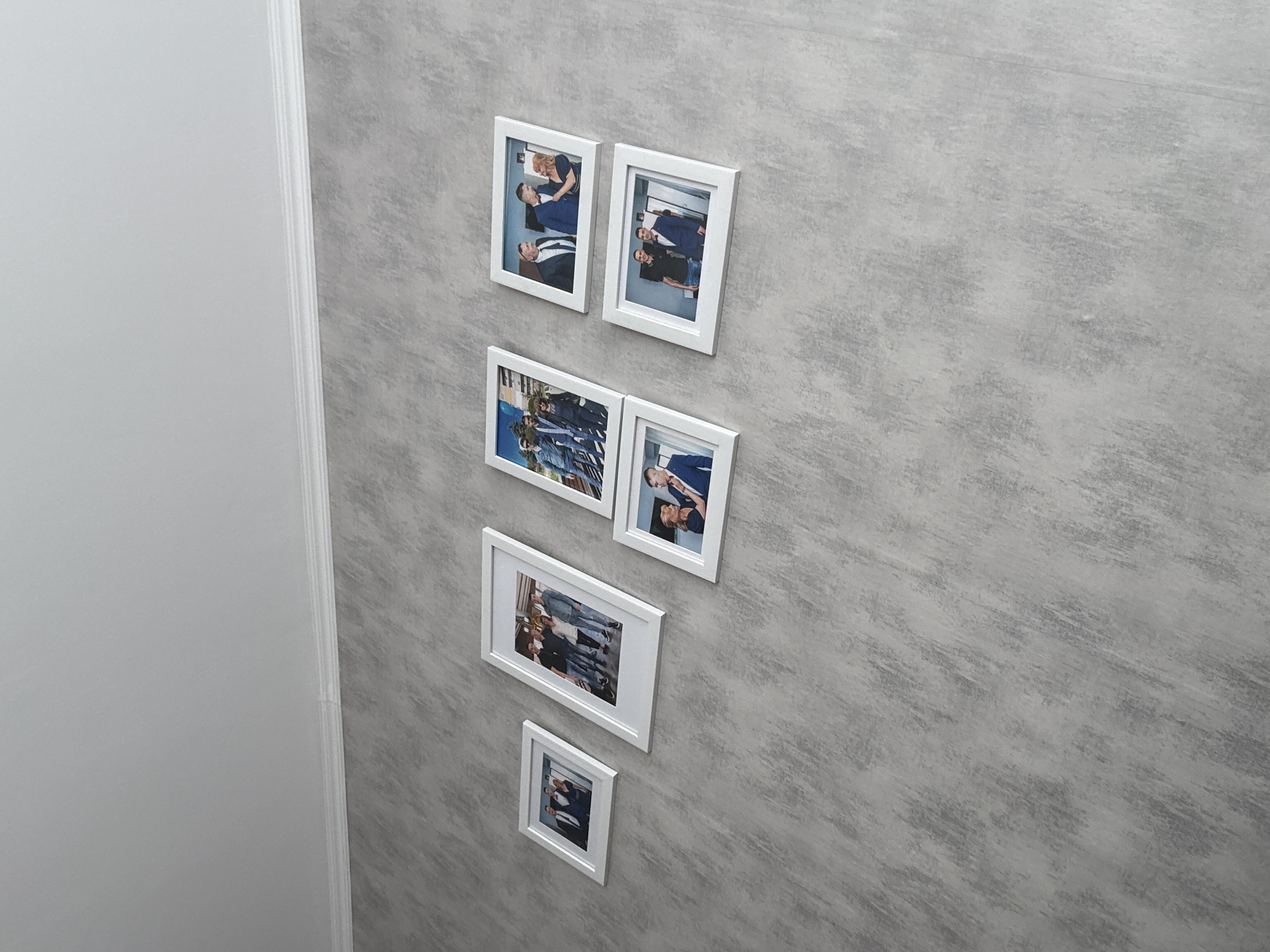 The image shows a vertical arrangement of five white-framed photographs on a gray wall with a textured finish, located beside a white wall in a room. The photographs appear to depict people, possibly in a formal or professional context. The overall scene is simple and domestic, suggesting a personal space such as a home or office. There is no indication of the location being in the city of Lovech, Bulgaria, or any other specific location.
