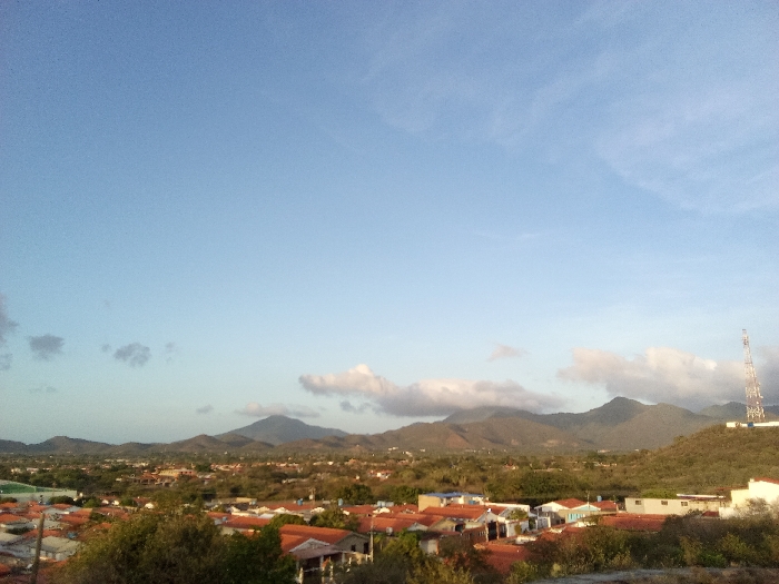 The image displays an elevated panoramic view over the city of Juan Griego, Venezuela. The scene is illuminated by soft, diffuse natural light, suggesting either early morning or late afternoon. The upper two-thirds of the frame consists of a clear, light blue sky with scattered white cumulus and wispy cirrus clouds.
In the lower third of the image, the foreground comprises dense, dark green foliage and some lighter-colored dry scrub, indicating an elevated observation point. Directly below, the midground is dominated by a residential area with numerous low-rise buildings. These structures predominantly feature reddish-orange tiled roofs, with some lighter-colored buildings also present. Green vegetation is interspersed among the dwellings.
The background reveals a range of dark green, vegetated mountains stretching across the horizon, with several peaks partially obscured by clouds. On a hill to the far right, a tall, lattice-structure communication tower is visible, extending into the sky. No individuals, specific actions, or interactions are discernible.