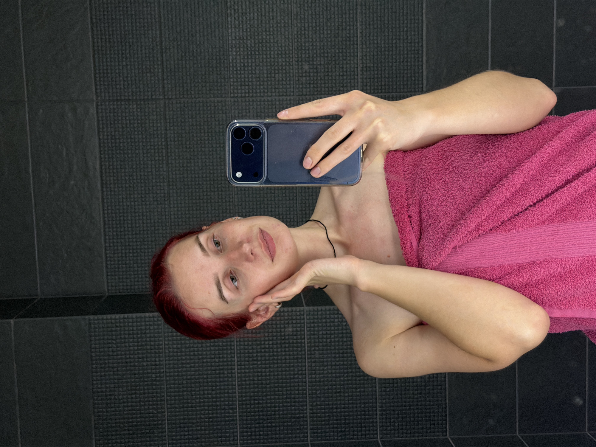 A young woman with bright red hair, wearing a pink towel, is taking a selfie in a bathroom. The background is tiled with dark gray square tiles. She is holding a dark blue phone in her right hand, with her left hand touching her cheek. She is looking at the phone.