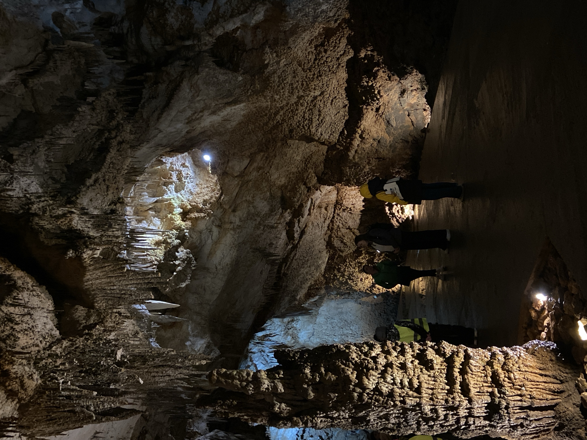 The image shows an interior view of Lipa Cave in Lipa Dobrska, Montenegro. Three individuals are positioned within the cave's illuminated interior, likely on a guided tour. The cave features formations such as stalactites and stalagmites, which are variably illuminated, highlighting their texture. The environment is primarily dark and cavernous, typical of cave systems.
