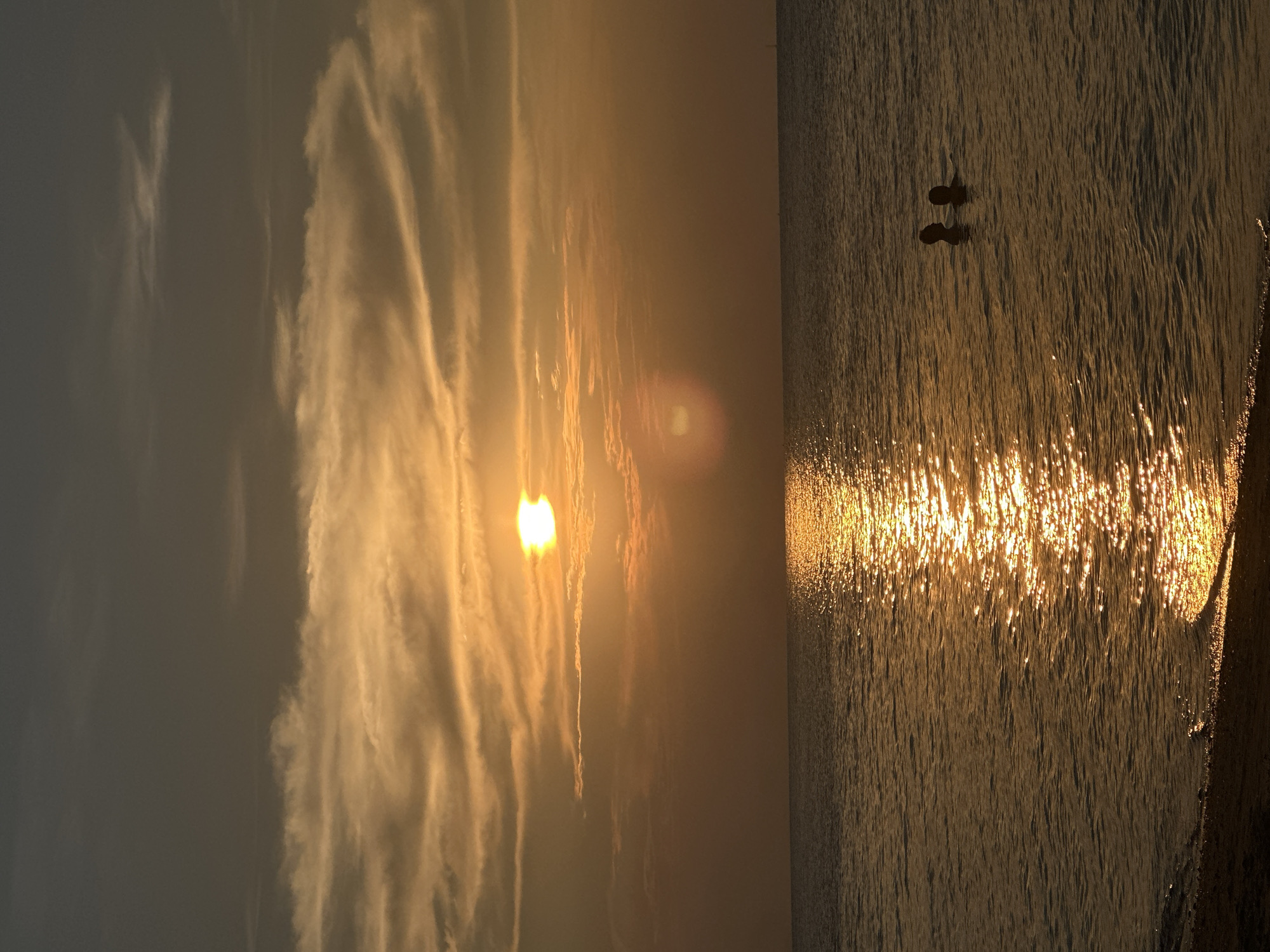 The image depicts a serene sunset or sunrise scene over a body of water, likely the sea, at Baan Tai, Thailand. The sun, a bright golden orb, is low on the left side of the horizon, casting a brilliant, shimmering reflection across the water towards the right side of the frame.
Two silhouetted figures, appearing to be people, are visible standing in the water on the right. Their dark forms are starkly contrasted against the intense golden light reflecting on the water's surface. They appear to be standing relatively still, possibly observing the scenic view.
The sky, particularly in the upper left portion, features wispy, elongated clouds that are illuminated in warm orange and yellow tones by the low sun, creating dynamic patterns. Away from the sun's direct glow, the sky transitions into a darker, muted grey-blue. The surface of the water is textured with small ripples and waves, which intensely catch and scatter the sunlight, forming a glittering pathway of gold extending towards the viewer. The overall atmosphere is one of calm and natural beauty, characterized by dramatic backlighting and warm hues. There is no visible text in the image.