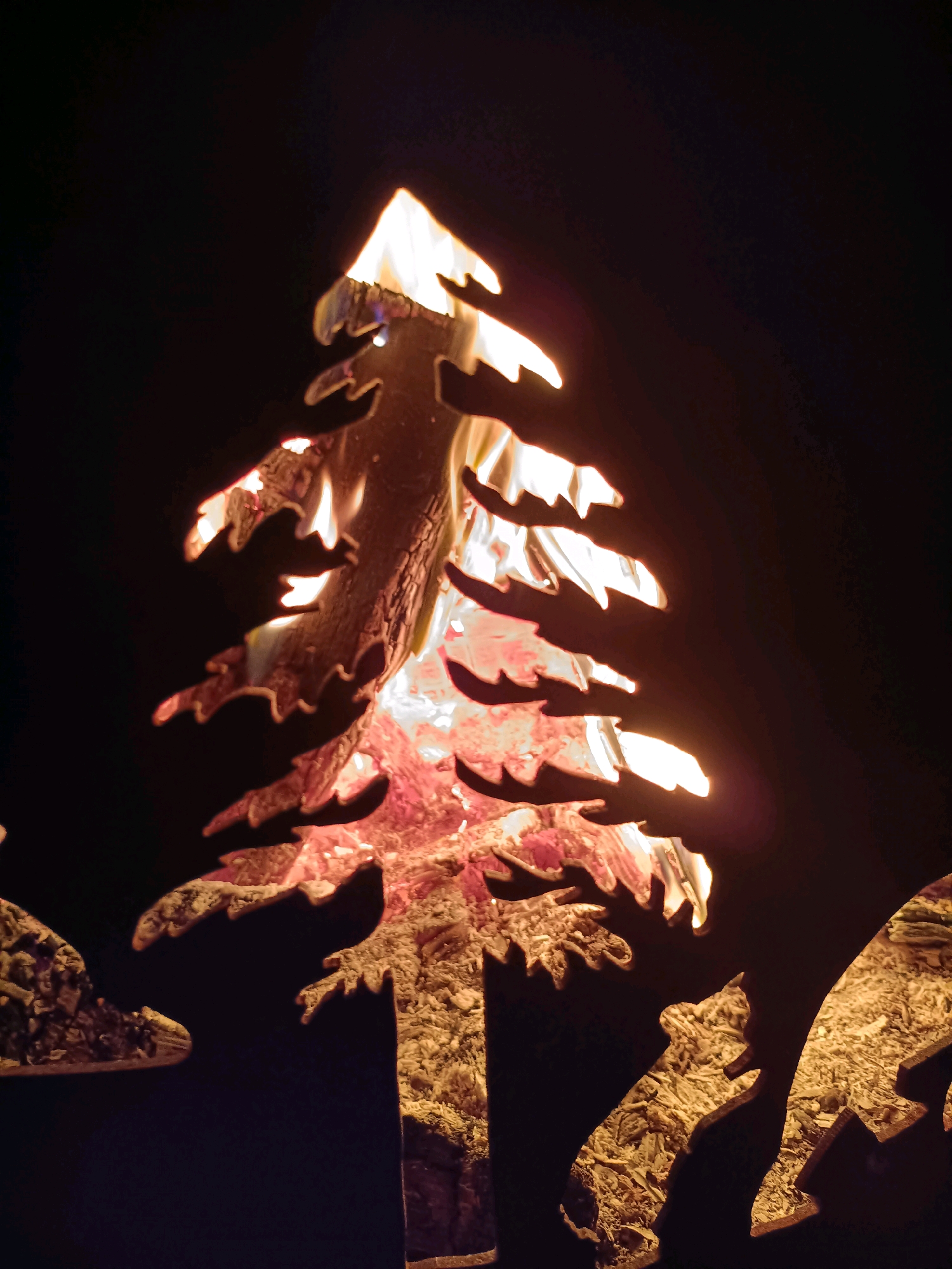 The image shows a metal cutout of a tree, with flames from a fire visible through the cutout. The fire illuminates the edges of the metal tree and casts shadows. In the foreground, there are other metal cutouts of animals, possibly a moose and a fox, also silhouetted against the fire. The ground beneath the fire appears to be covered in wood shavings or kindling. The background is dark, suggesting this scene takes place at night. The location is stated to be in the city of Tiraspol, Moldova.