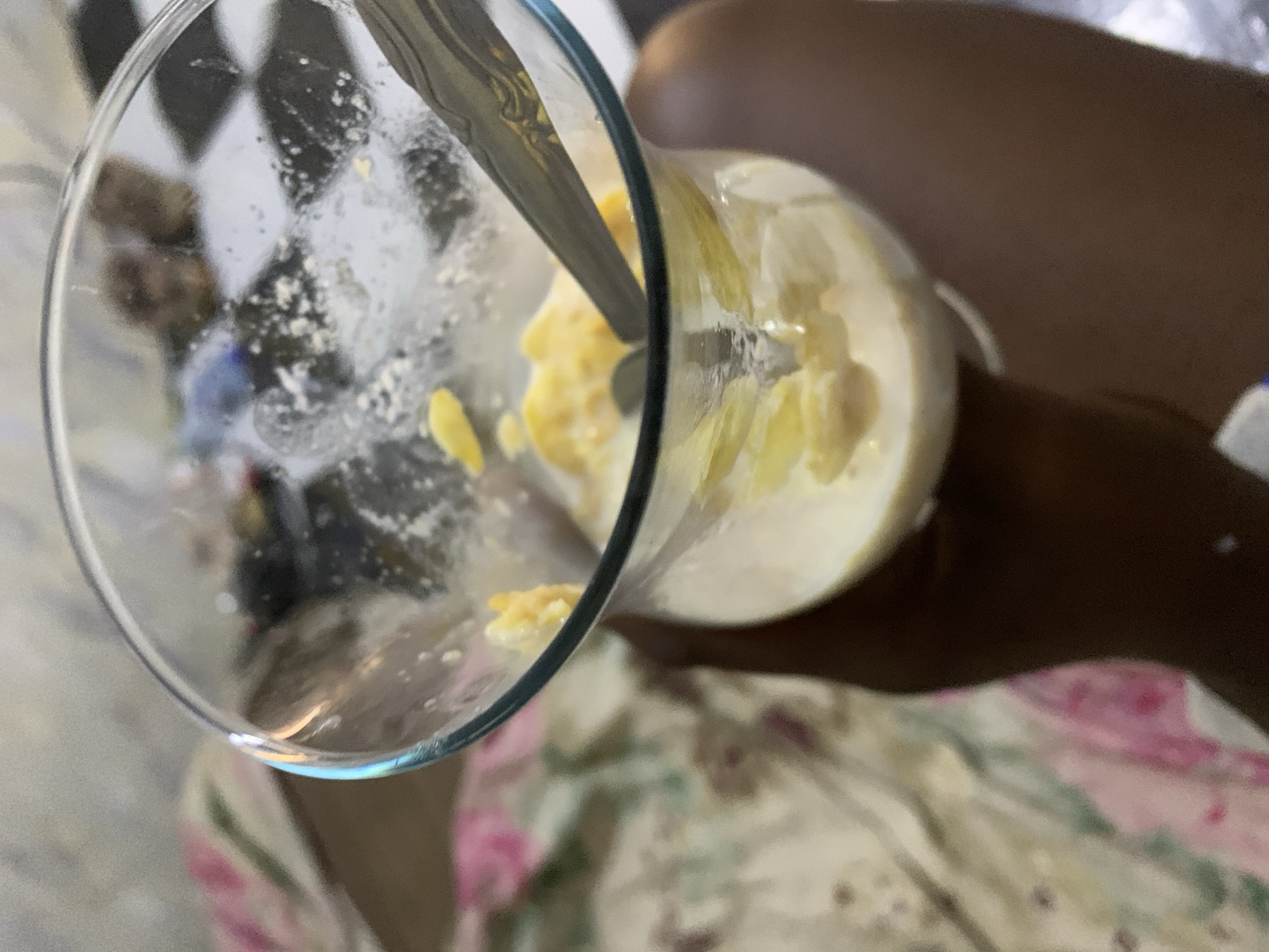 A glass of yellow and white smoothie-like liquid is being held by a dark-skinned individual wearing a floral patterned garment. A metal spoon is inside the glass. The background includes a checkered pattern, likely a wall or other surface. The location is unknown.
