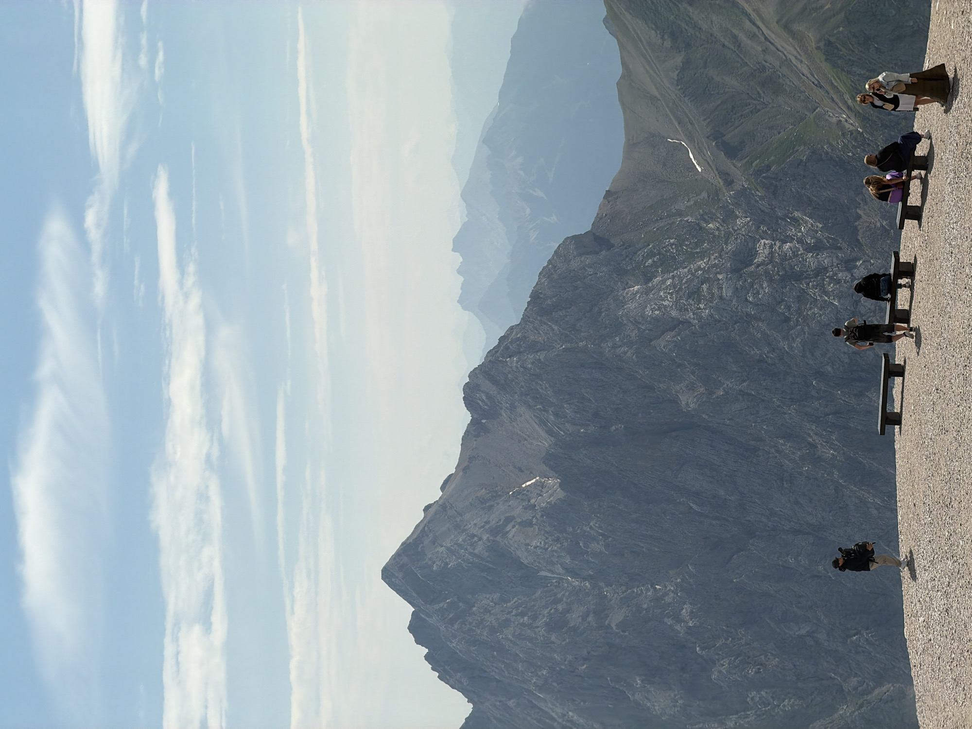 This media file depicts a panoramic view of a mountainous landscape, likely taken from a high vantage point. The scene is oriented vertically, with the sky and distant mountains filling the upper portion and a close-up of a rocky mountainside dominating the lower half. Several people are visible, positioned on a path or viewing area along the edge of the mountain.
In the foreground, a person with a backpack is seen from behind, facing away from the camera and towards the vast mountain range. Further along the path, several other individuals are either sitting on benches or standing, appearing to admire the scenery. One group of three women is posed together, likely for a photograph. Another group of two is seen sitting on a bench, and a solitary figure is also visible on a bench. The attire of the people suggests a casual, outdoor setting, possibly a tourist destination.
The mountains are rugged and layered, with slopes covered in a mix of rock and sparse vegetation. Faint white lines, possibly snow patches or streaks of light, are visible on some of the slopes. The sky is bright blue with wispy white clouds streaking across it, indicating fair weather and likely daytime. The lighting suggests it could be mid-morning or afternoon, with the sun casting shadows.
The presence of benches and a paved walkway suggests a developed tourist area, perhaps a viewpoint or a trail leading to one. The overall atmosphere appears serene and awe-inspiring, with people enjoying the dramatic natural beauty of the location. The location is identified as Garmisch-Partenkirchen, Germany, which is known for its alpine scenery. No text is visible in the image.