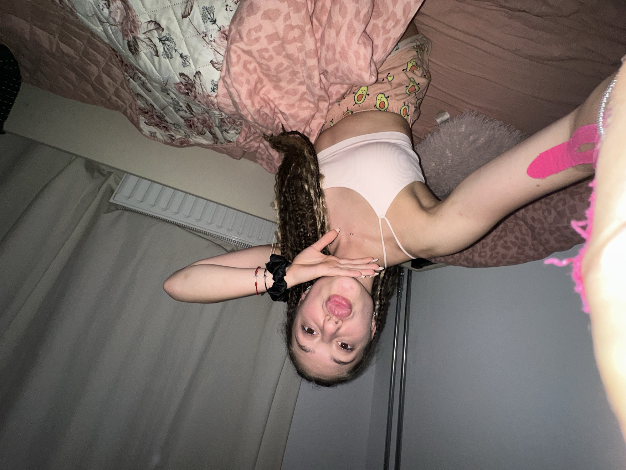 A young woman with light brown curly hair pulled into braids is lying on a bed, taking a selfie. She is wearing a light pink crop top and patterned pink shorts with avocados on them. Her left arm is extended toward the camera, and her right hand is resting on her chin. She has a playful expression, with her lips pursed. The bed is covered in pink and floral bedding. The background shows a wall, a window with curtains, and a radiator. The scene appears to be an indoor bedroom. There is no specific indication of the location being in the city of Sofia, Bulgaria.