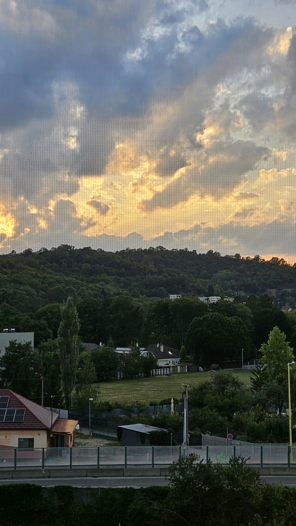 This image depicts a view of a forested hillside with scattered houses and a road in the foreground, set against a dramatic cloudy sky at sunset. The sky is filled with large, textured clouds, some of which are illuminated by the setting sun with warm golden and orange hues, while others are a darker blue. The forested hill rises in the mid-ground, with the dense green foliage partially obscuring buildings nestled within. In the foreground, a house with a red tiled roof and solar panels is visible on the left, alongside a road or highway bordered by a green fence. The scene appears to be a suburban or semi-rural area. The image has a slightly hazy or diffused quality, possibly due to it being taken through a screen. The composition suggests a tranquil, late afternoon or early evening setting. The specific location within the city of Prešov, Slovakia, cannot be definitively identified from the visual information provided alone.