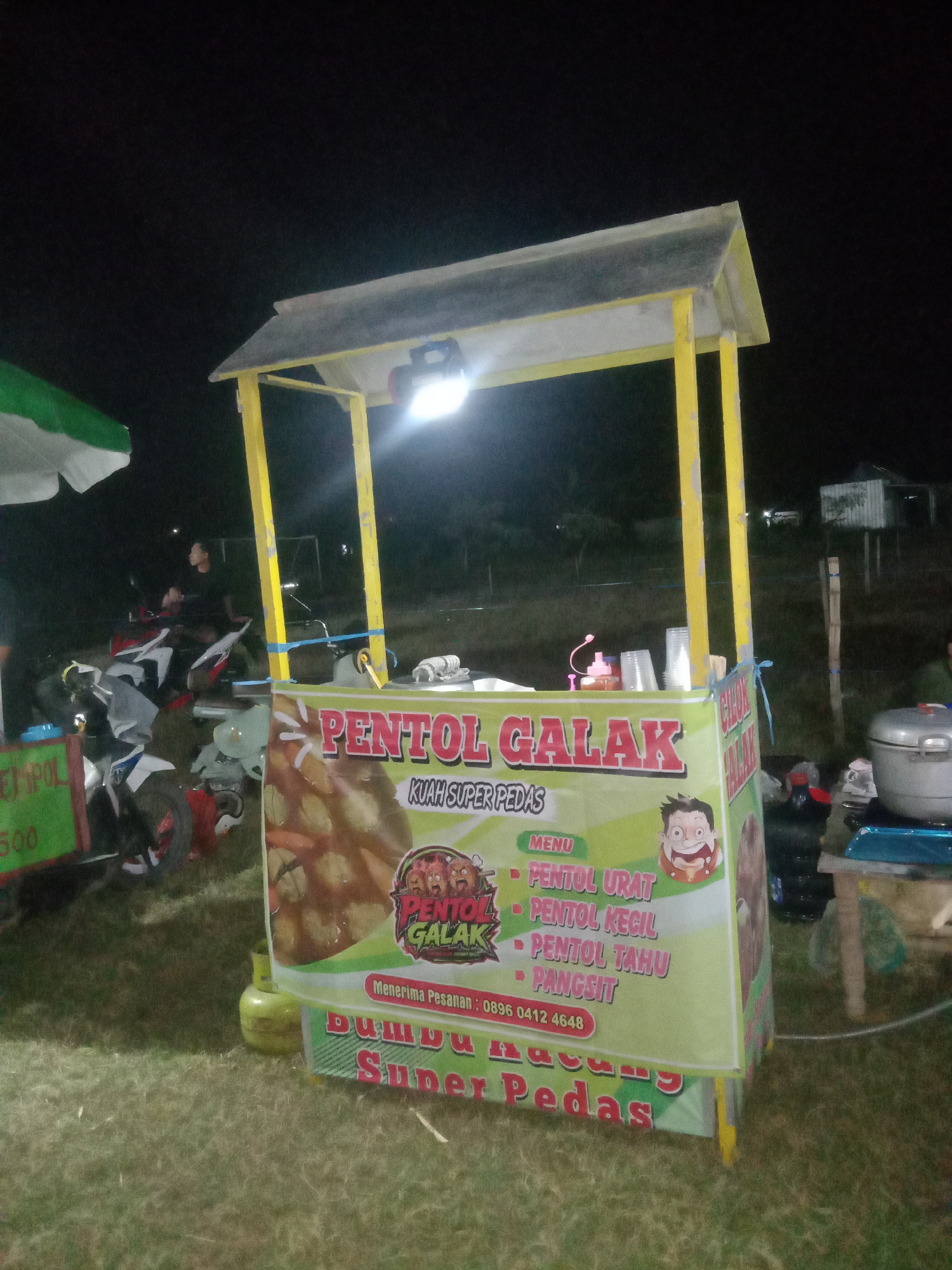 The image captures a food stall at night in what appears to be an outdoor, possibly grassy area. The stall is illuminated by a bright light hanging from its yellow roof. Prominently displayed on the stall is a banner advertising "PENTOL GALAK" (Spicy Pentol), with the subheading "KUAH SUPER PEDAS" (Super Spicy Gravy). The banner also lists menu items: "PENTOL URAT" (Vein Pentol), "PENTOL KECIL" (Small Pentol), "PENTOL TAHU" (Tofu Pentol), and "PANGSIT" (Wonton). A phone number, "0896 0412 4648", is also visible for orders. Below this, another banner reads "BUMBU KACANG SUPER PEDAS" (Super Spicy Peanut Sauce).
In the background, several motorcycles are parked, and a person can be seen sitting on one of them. To the left of the stall, a portion of a green and white umbrella is visible. To the right, a large silver pot is placed on a table, suggesting food preparation or service. The overall setting suggests a food vendor operating during an evening event or in a popular nighttime gathering spot. The mention of Jumput, Indonesia, in the prompt suggests this scene is located within that city.
