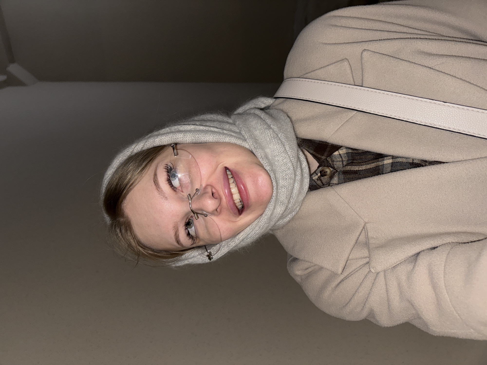 A young woman is captured in a close-up shot, looking upwards and smiling broadly. She has light-colored hair, partially visible beneath a light beige or cream-colored hooded scarf or cowl that frames her face. She wears rimless glasses with delicate metal frames, reflecting light. Her eyes are open wide, looking slightly to her left, with prominent eyelashes. Her smile reveals her upper teeth.
Over her plaid shirt, which features shades of brown, cream, and grey, she wears a light beige or cream-colored wool coat. A white, textured shoulder strap from a bag runs across her chest. The background is uniformly dark and out of focus, suggesting an indoor setting with low ambient light, or a close-up shot against a plain wall. The direct lighting on her face and attire suggests a flash or a strong single light source. Her expression is cheerful and engaging. There is no visible text in the image.
