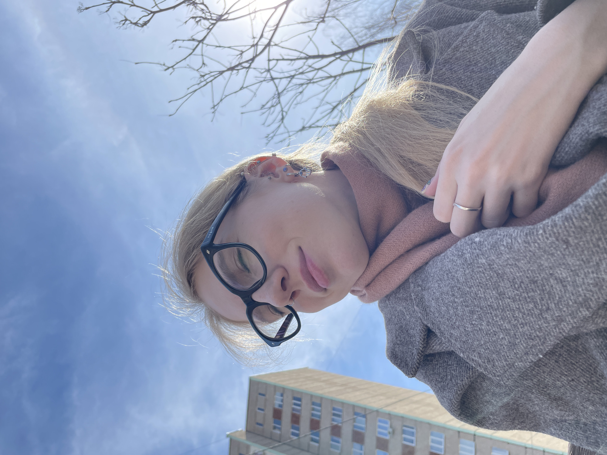 A selfie, taken from a low angle, shows a woman with blonde hair, glasses, and a light pink scarf wearing a gray coat. The sky is blue with bare tree branches in the upper part of the frame. A light-colored high-rise building with repeating window patterns is visible in the lower portion of the frame. The building is located in Izhevsk, Russia.