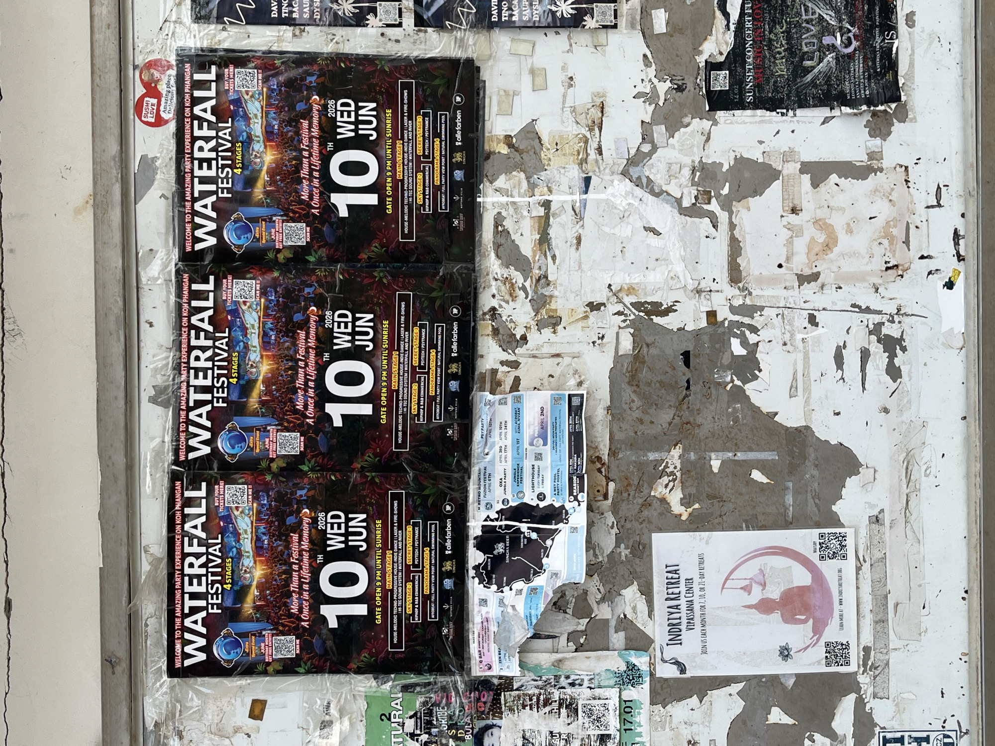 This image captures a weathered public announcement board or wall in ตำบลเกาะพะงัน, Thailand, covered with multiple overlapping event posters and the remnants of torn flyers. Dominating the upper portion are three identical "Waterfall Festival" posters, each featuring a black background adorned with lush, red-leafed tropical foliage, a vibrant festival crowd, and an illuminated stage. These posters advertise an event on "10 TH WED JUN 2026," with "GATE OPEN 9 PM UNTIL SUNRISE," promising an "Amazing Party Experience" with "4 Stages" (Main Stage 1: House/Techno, Sky Stage 2: Hiphop/R&B, Secret Stage 3: Psytech/Psytrance, Panorama Stage 4: Afrobeat). QR codes are present for ticket purchases and a "June Birthday Month" free entry offer. Above the rightmost festival poster, a small red heart-shaped sticker reads "SUSHI LOVE Amazing place Delicious food."
Below the main festival posters, the wall reveals extensively peeled paint and numerous fragmented event advertisements. A prominent, partially obscured black map outline of Koh Phangan, labeled "SINGHA BEER," is visible in the center-left mid-section, surrounded by other smaller flyers detailing various parties and events, including "RETRO MOUNTAIN FUSION FESTIVAL APRIL 6TH," "OXL JUNGLE PARTY APRIL 1TH APRIL 24TH," "PSYPARTY APRIL 13TH," and "LIGHTHOUSE EVERY WEDNESDAY LIVE MUSIC APRIL 2ND."
In the bottom left quadrant, a white poster for "INDRIYA RETREAT VIPASSANA CENTER" invites participation in "7, 10, OR 21-DAY RETREATS," illustrated with a pink Buddha figure set within a crescent shape, a lotus flower, and hanging lamps, alongside a QR code and website address. The bottom right displays a partially visible dark-toned poster, possibly for a "SUNSET CONCERT" or "MUSIC IN LOVE" event, with the word "LOVE" prominently displayed and a date "14.02" visible. The wall itself is light-colored, showing significant material loss from peeling paint, revealing darker underlayers, and featuring a horizontal metal or wooden beam running across its upper section, just below a visible horizontal crack in the painted surface above the posters.