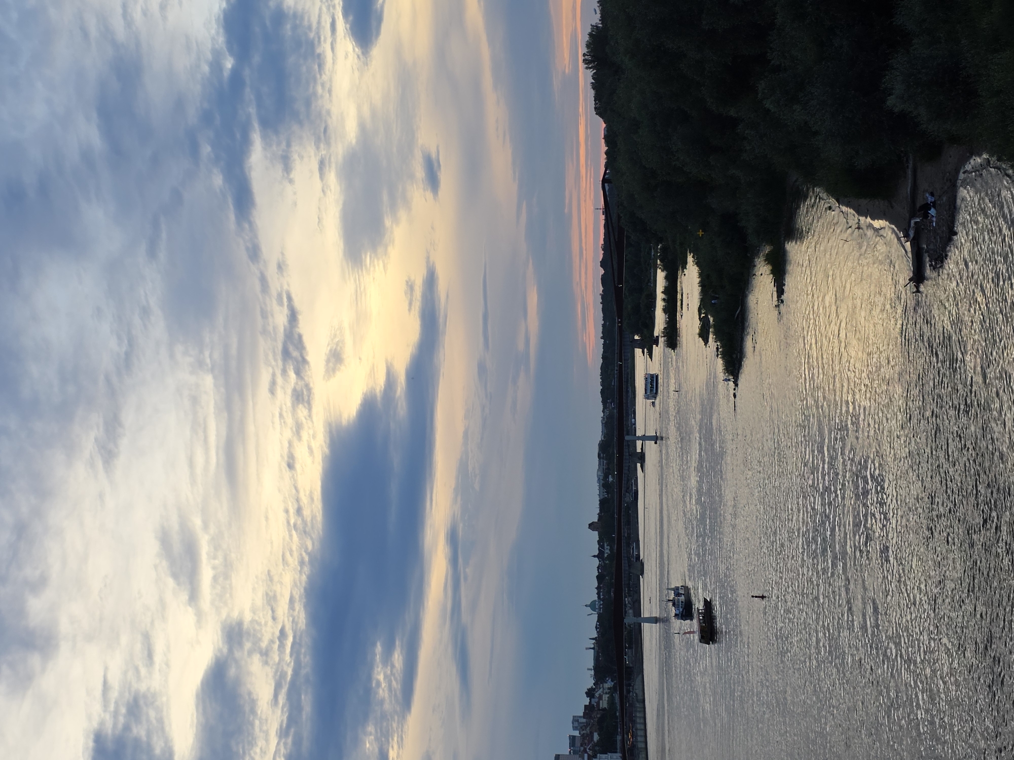 The image depicts a panoramic view of the Vistula River in Warsaw, Poland, during dusk. The sky is filled with dramatic clouds tinged with yellow and orange hues from the setting sun, contrasting with patches of blue and grey. A long bridge spans across the river in the mid-ground, with several boats of various sizes navigating the water. On the left bank, the cityscape of Warsaw is visible with buildings and structures, including what appears to be domes and towers. The right bank is densely covered with trees and vegetation, forming a dark, lush shoreline. The water reflects the colors of the sky and the surrounding environment, creating a serene and atmospheric scene. A few individuals are visible on the sandy shore of the right bank, seemingly relaxing or enjoying the evening.