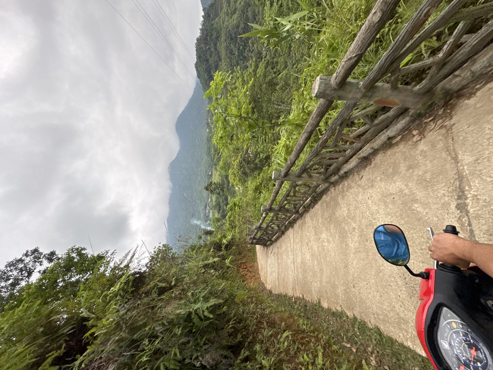 A first-person perspective shot shows a person riding a red scooter along a paved path in Pù Luông Commune, Vietnam. The path curves to the left, bordered on the left by a steep, vegetated hillside and on the right by a rustic wooden fence. Lush green foliage covers the hillside and the landscape beyond the fence. In the distance, rolling hills covered in dense forest rise under a cloudy sky. Power lines are visible in the upper right corner of the frame. The rider's left hand is on the handlebar, and their right hand is gripping the throttle. The rider is wearing a blue shirt, reflected in the scooter's rearview mirror. Smoke is visible rising from a small settlement in the valley in the distance.