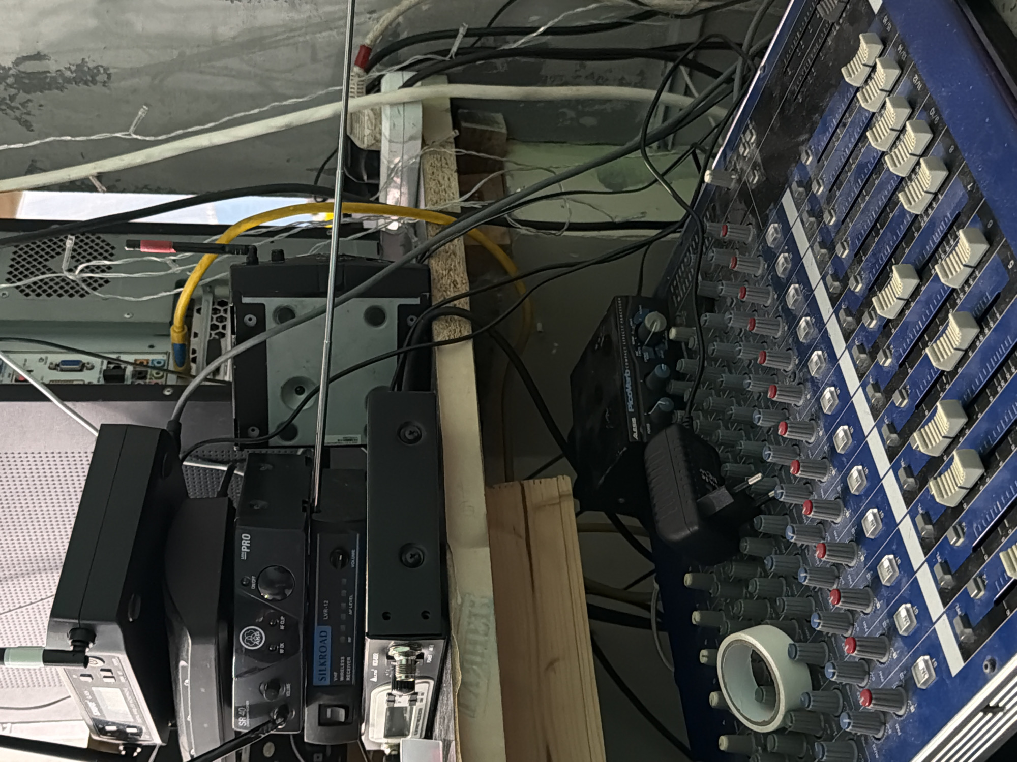 The image shows a cluttered technical setup, likely for audio production. On the right, a blue mixing console with numerous knobs and sliders dominates the view. To the left, various black audio receivers and processors are stacked together. Visible labels on these devices include "SILKROAD LVR-12" and "AKG SR40". There is also an "ALESIS PicoVerb PROCESSOR" partially visible. Wires and cables are intertwined throughout the setup, suggesting a complex and potentially messy workspace. A section of a computer with network ports can be seen at the top left of the frame. There are no people visible in the image. There is no explicit confirmation of the location in Perm, Russia.