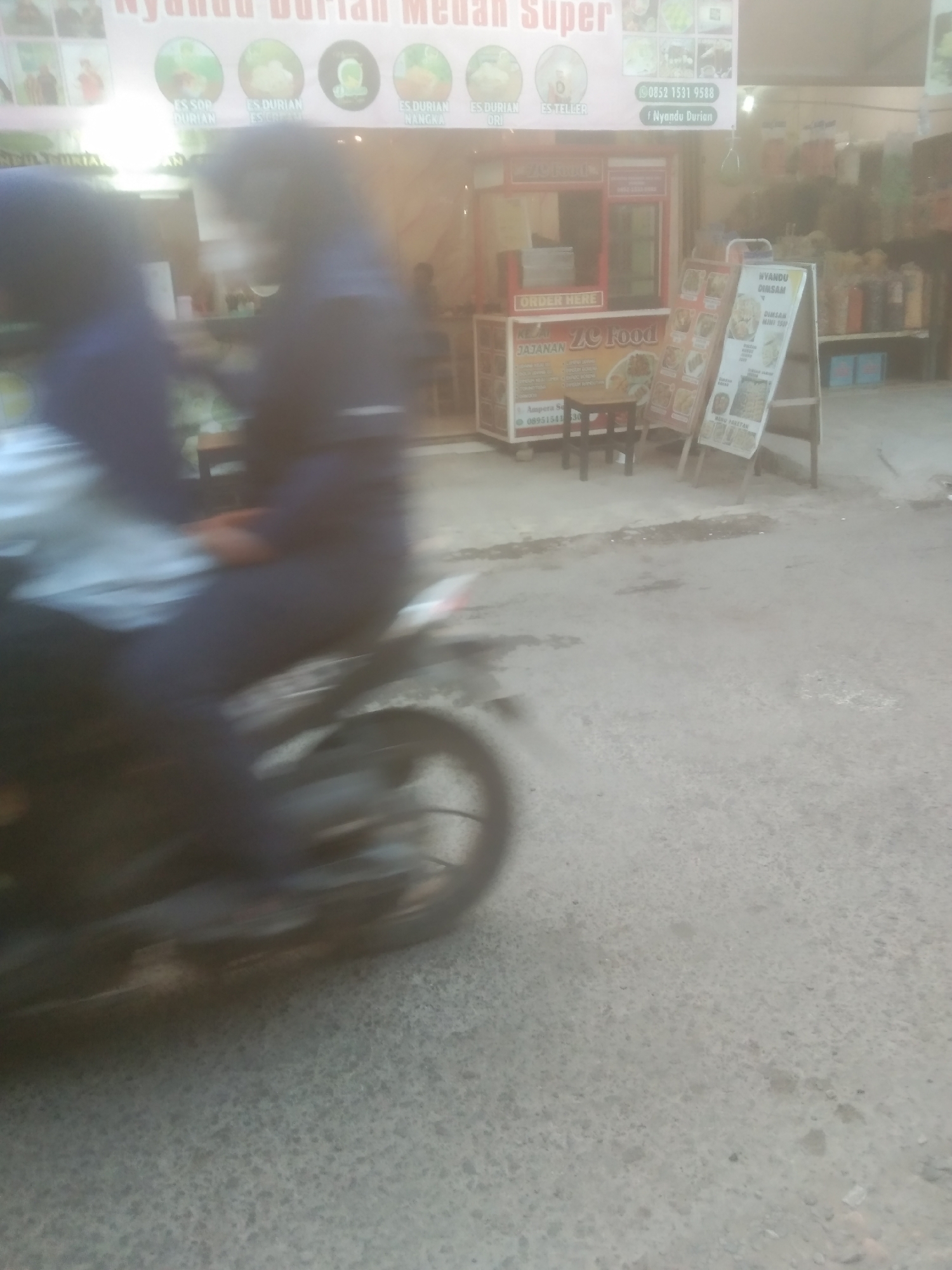 This is a motion-blurred, medium-distance, eye-level, outdoor photograph taken on a street in Subang, Indonesia. The foreground is dominated by a motorcycle moving from left to right, carrying two individuals. The driver, in the front, is wearing a dark blue long-sleeved shirt, and the passenger, behind, is wearing a dark blue hijab and a dark blue long-sleeved shirt. The motorcycle is black and appears to have a white object on its rear. The background features a small food stall with a red and white facade and various signs advertising food and drinks. One sign in the background appears to say "Nyandu Durian Medan Super." Adjacent to the food stall is a menu board on an easel advertising food items, and another board with the text "NYANDU" and "DURIAN." The pavement is rough and grey, with some debris scattered on it. The overall lighting suggests it is daytime, but the motion blur obscures many finer details.