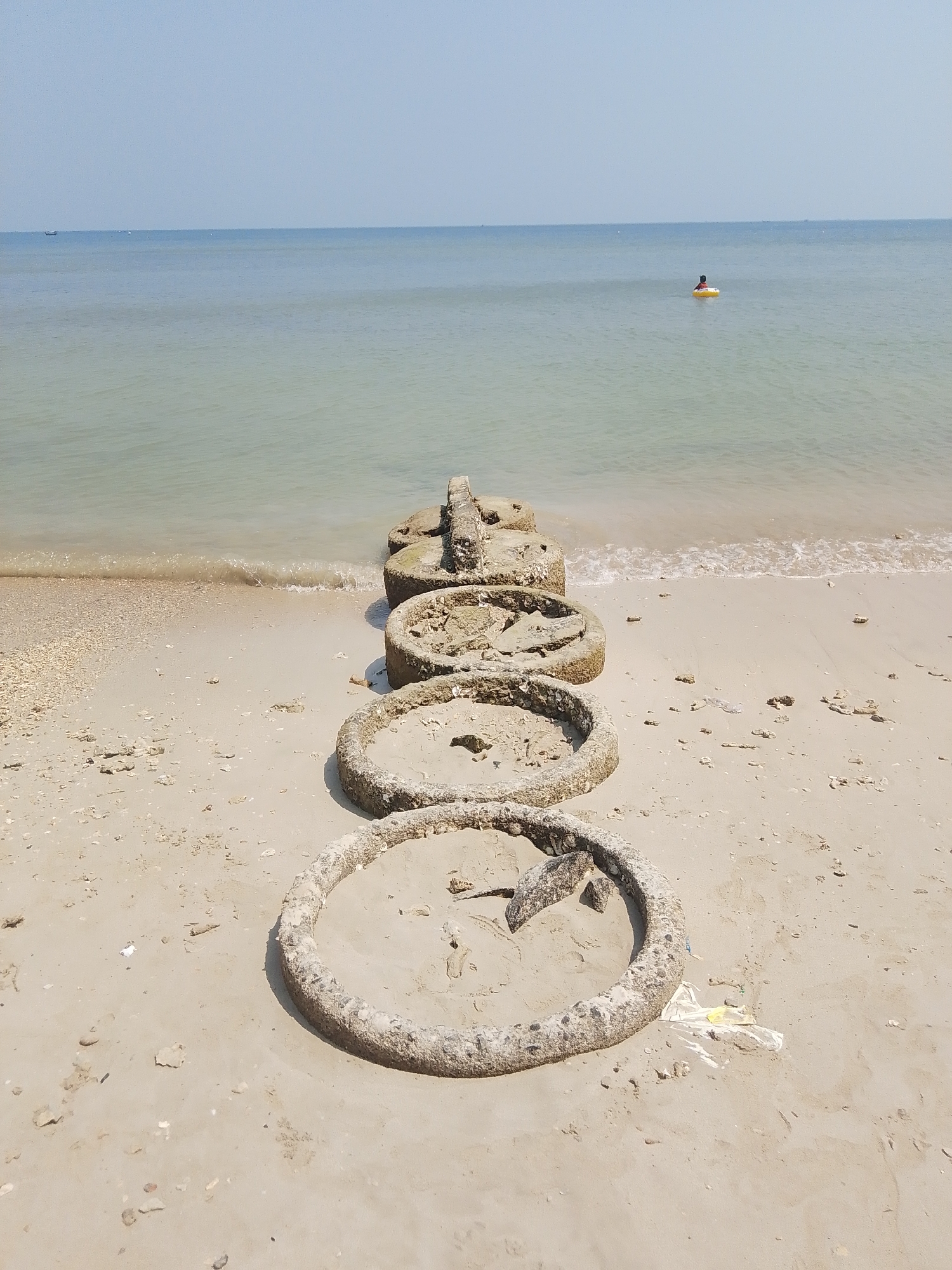 The image depicts a tranquil beach scene in Tegalsambi, Indonesia. In the foreground, several weathered concrete rings lie partially buried in the sand, arranged in a staggered line perpendicular to the shoreline. These appear to be remnants of some past construction or debris. The sand is light brown, scattered with small shells and pebbles. Gentle waves lap at the shore, creating a narrow strip of wet sand.
In the background, the calm, light blue sea stretches to the horizon under a clear sky. A lone figure is visible in the water, sitting on a yellow inflatable ring, indicating recreational activity. A small boat can be seen in the far distance on the left side of the image. The overall atmosphere is peaceful and serene.