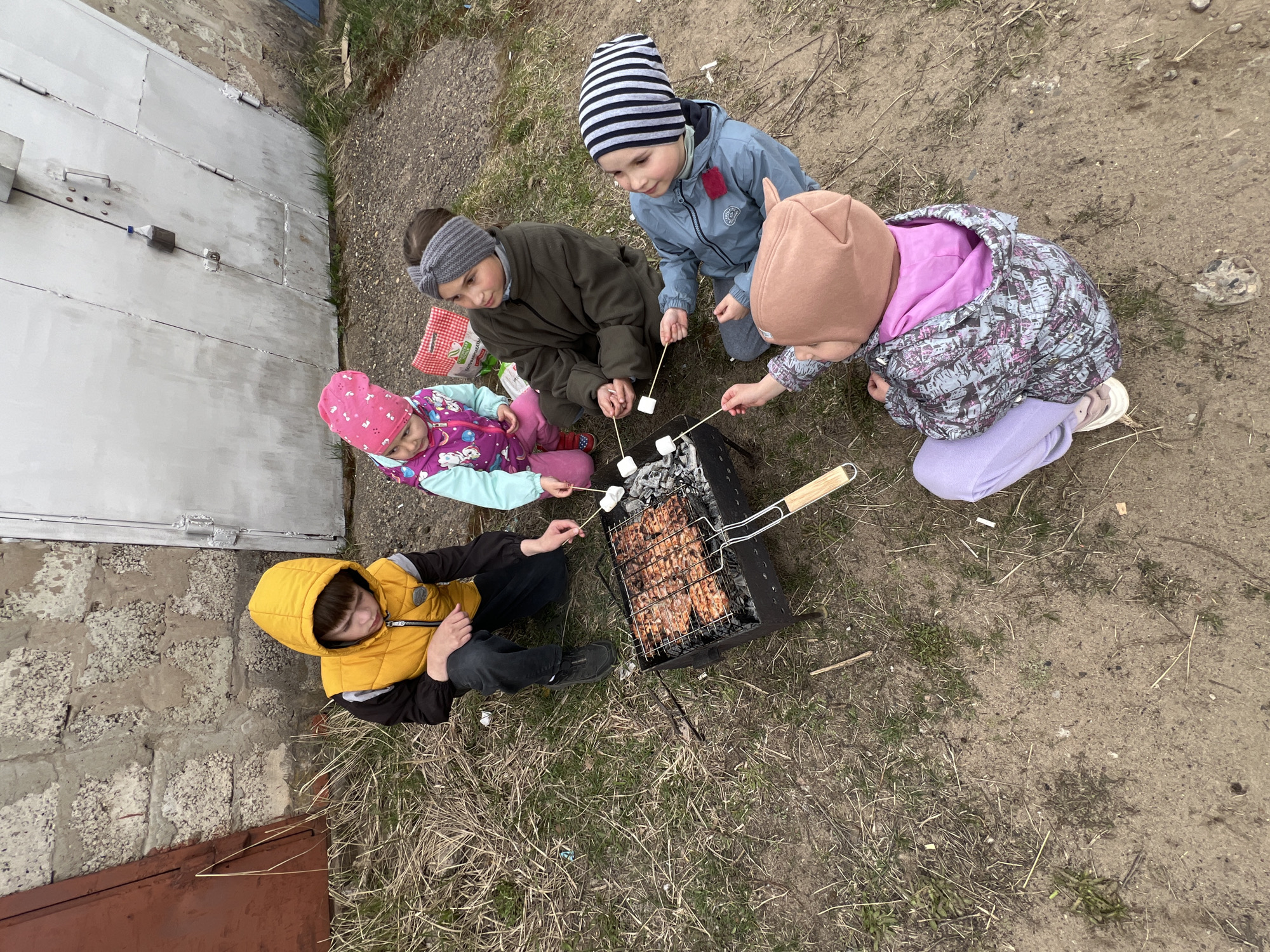 Five children are gathered around a charcoal grill. The grill is cooking meat. Four of the children hold sticks with marshmallows over the grill. The children are wearing various hats and jackets. One child is wearing a yellow vest. The ground is dirt and sparse grass. A grey garage door and brick wall are in the background. The setting is Noviy, Russia.