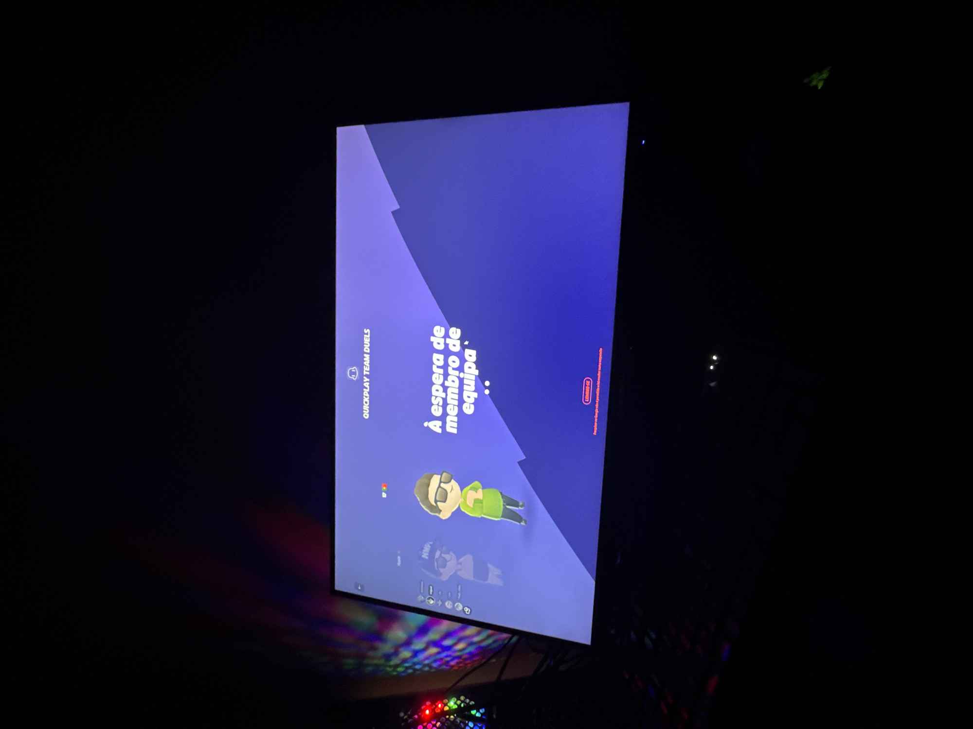 The image shows a television screen displaying a waiting screen for the video game "Multiversus." The screen reads "À espera de membro de equipa," which translates to "Waiting for team member" in Portuguese. The game mode is "Quickplay Team Duels." A character avatar is visible on the screen. The location is likely inside a home, as indicated by the dark environment, and a partially visible keyboard with colorful backlighting. The geographic location is not evident from the image.