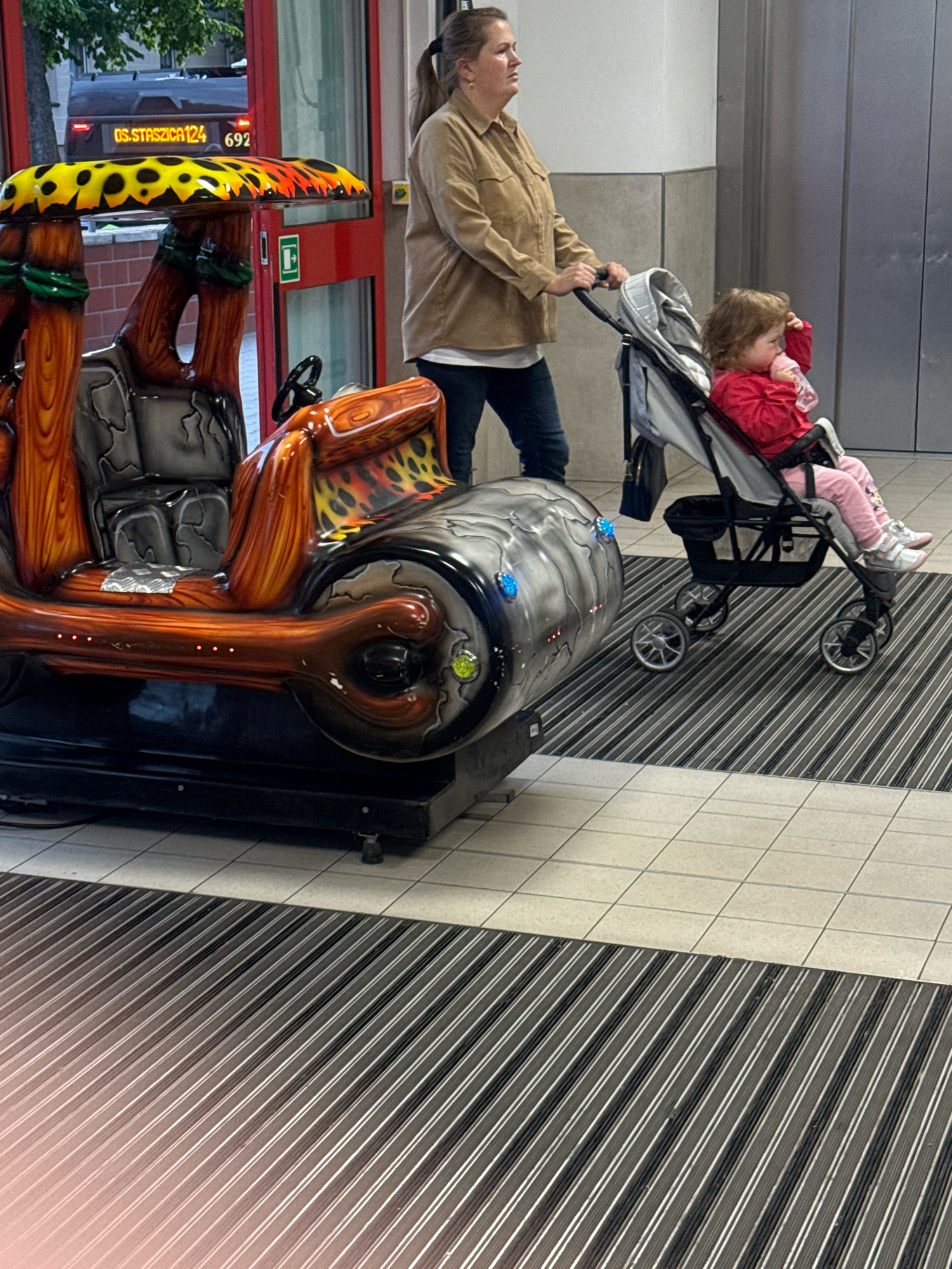 A woman in a brown jacket and jeans pushes a stroller with a toddler in a red top and pink pants. The toddler is drinking from a bottle. The stroller is gray and has four wheels. They are on a striped mat in front of a building with a glass door. To the left is a colorful children's amusement ride shaped like a car with a stone-like design. The ride has a yellow and black spotted roof with red flames. A bus sign that reads "OS.STASZICA 124 692" is visible in the background through the glass door. The scene appears to be a public indoor space, possibly a shopping center or train station, in Gorzow, Poland.