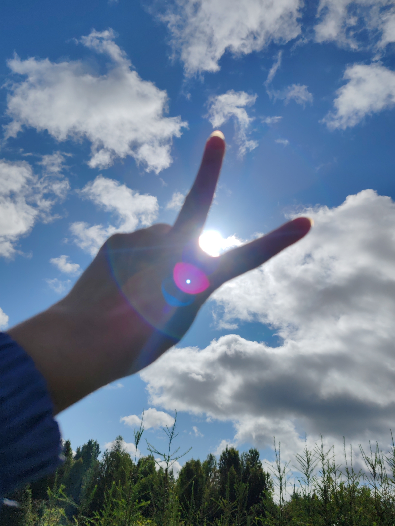 A hand is held up against a bright blue sky filled with white and gray clouds. The sun is behind the hand, creating lens flare with pink and blue hues. The hand is making a peace sign, with the index and middle fingers extended. The bottom of the frame shows a dark green line of trees. The image is taken outdoors, likely in a rural or natural setting. The location is not specified within the image itself.