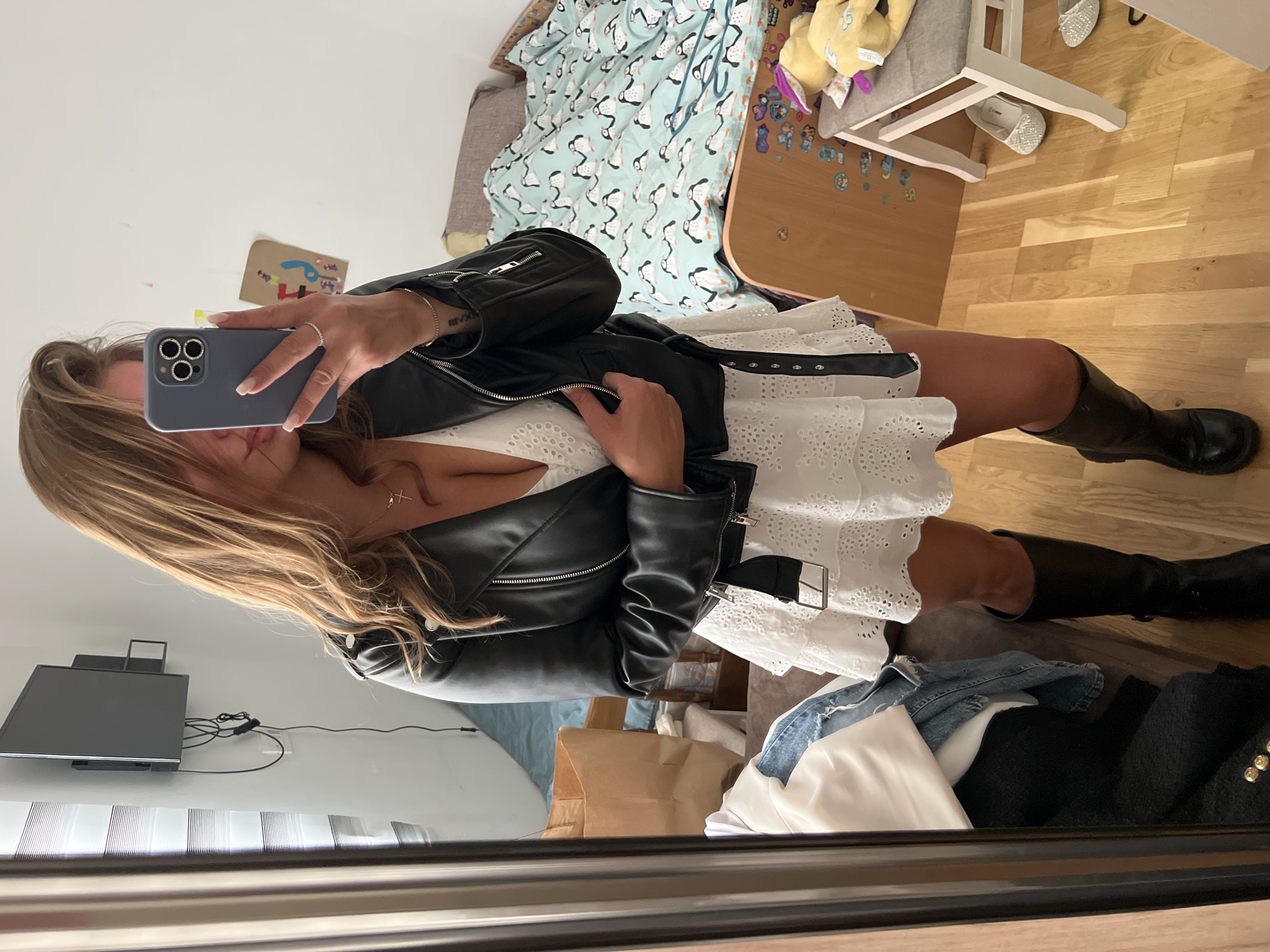 A mirror selfie shows a woman indoors, possibly in an apartment or hotel room in Budva, Montenegro. She holds a smartphone, obscuring her face, wearing a white eyelet dress, a black leather jacket, and black knee-high boots. The background shows a bed with penguin-patterned linens, a child's table and chair, and clothing strewn on the floor. A television is mounted on the wall.