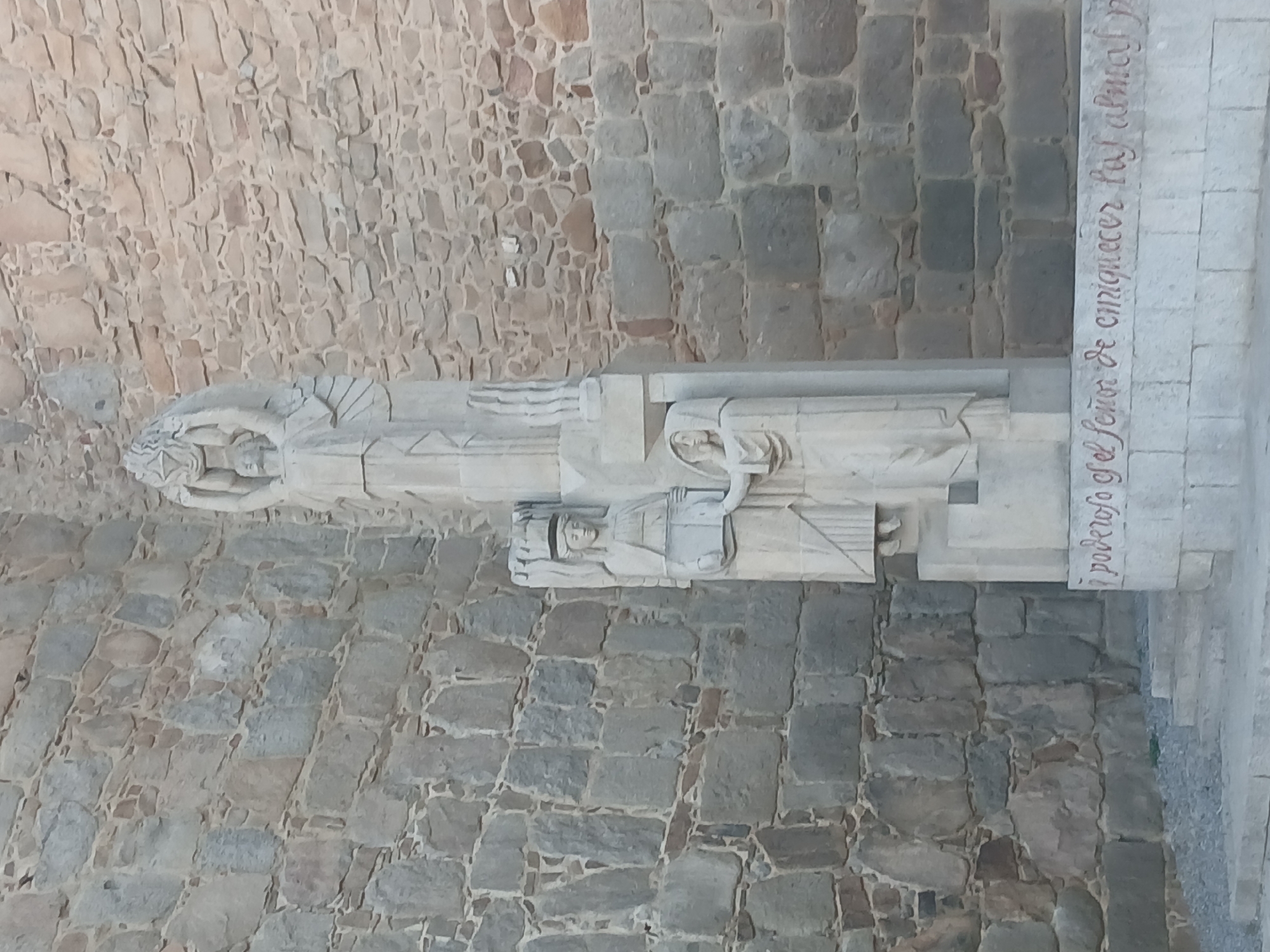 The image shows a stone sculpture in front of a stone wall in the city of Avila, Spain. The sculpture is a tall, slender monument composed of several figures. At the top, a large figure with outstretched arms appears to be holding a circular object or crown, with wings or decorative elements extending from its sides. Below this figure, a smaller figure stands with a headdress and holds an object that resembles a book or tablet. To the right of this figure, a third, smaller figure, draped in robes, extends a hand towards the second figure. The base of the sculpture has red lettering carved into it. The stone wall behind the sculpture is constructed from rough-hewn, irregular stones of varying sizes and colors, with visible mortar lines. The ground in front of the sculpture consists of a small set of stone steps leading up to the monument, with gravel and some sparse vegetation around the steps. The lighting suggests it is daytime, with the sun casting some subtle shadows.