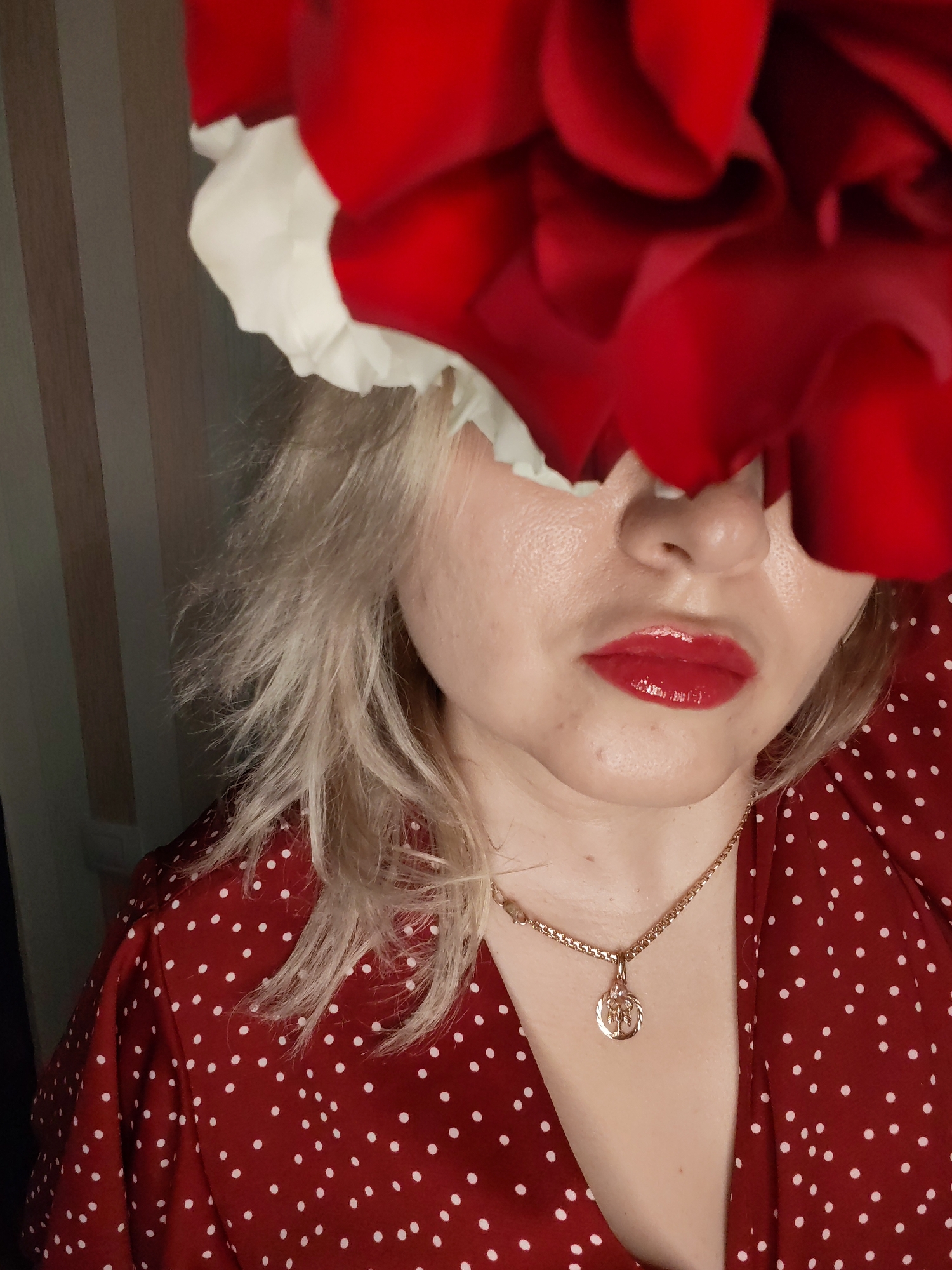 A close-up portrait features a woman with her upper face, including her eyes, obscured by a large cluster of vibrant red and white flowers. Her visible features include her nose, mouth, and chin. She has fair skin with some textural imperfections and wears glossy, bright red lipstick. Her blonde hair appears somewhat disheveled, framing her face and resting on her shoulders.
She is dressed in a red garment, likely a top or dress, patterned with numerous small white polka dots. Around her neck, she wears a gold-colored chain necklace with a small, round gold pendant that displays an intricate, possibly floral or symbolic, design.
The background consists of vertical stripes in varying shades of light brown or beige, indicating an indoor setting with a paneled or wallpapered wall. The lighting suggests an artificial source, casting highlights on her skin and the glossy lipstick. The woman is intentionally posing, using the flowers to create an artistic or enigmatic effect. There is no visible text in the image. The location context provided is Izhevsk, Russia.