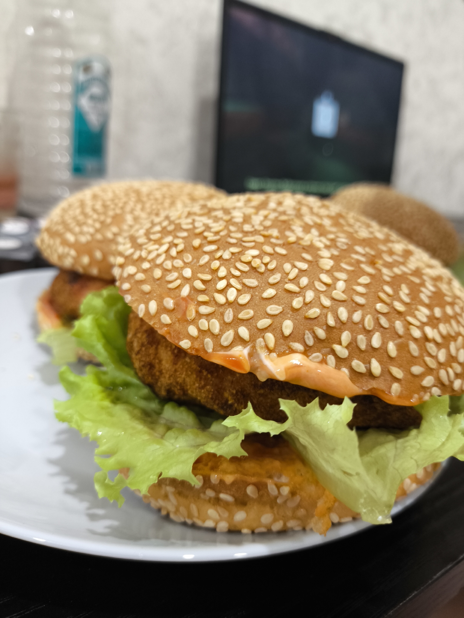 A close-up, side-view, eye-level shot shows two vegan burgers on a white plate. The burgers have sesame seed buns, lettuce, and what appears to be a vegan patty. The background is blurred and includes a water bottle and a laptop. The scene is indoors. There is no indication of the location in Penza, Russia.