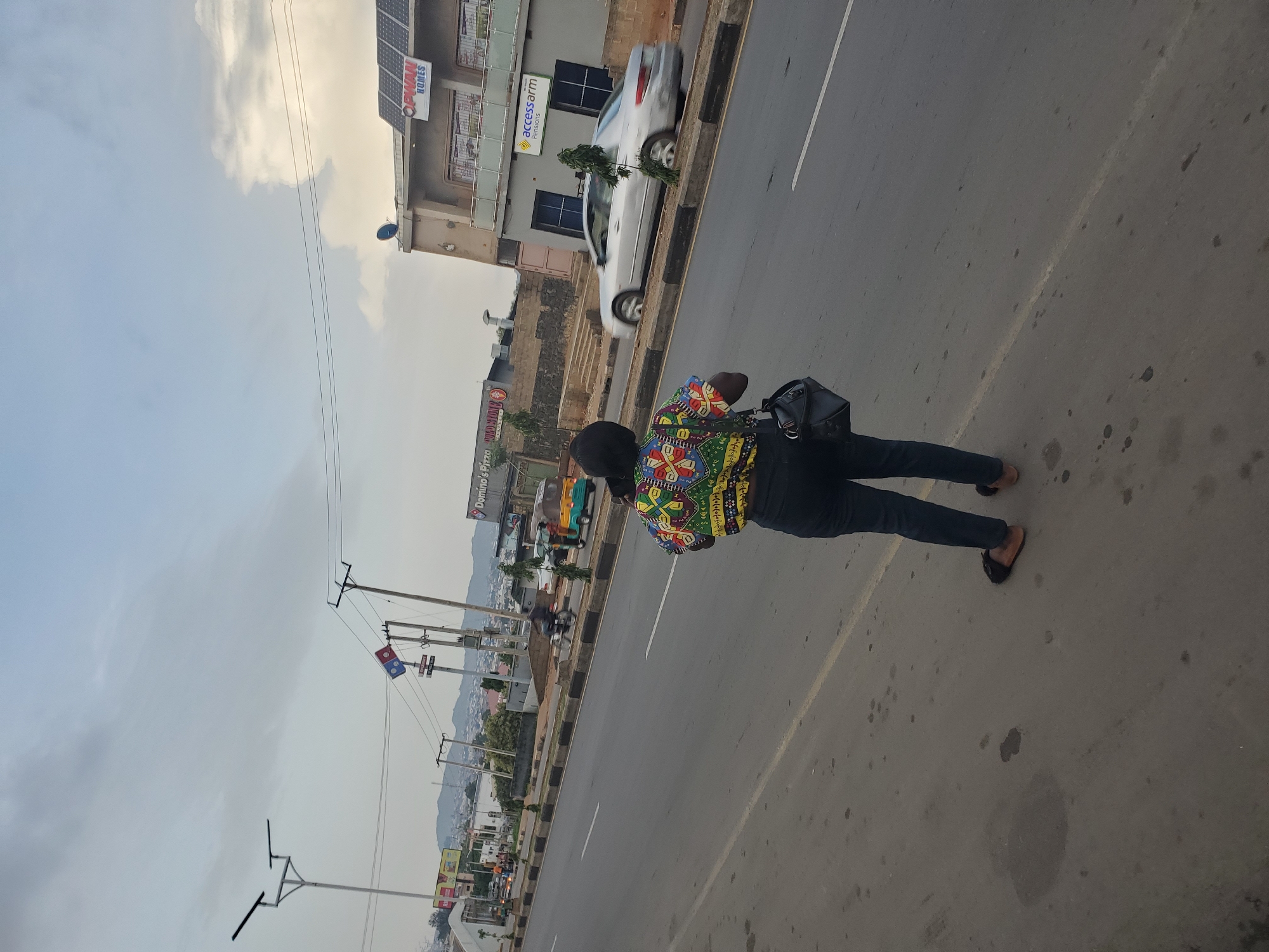 A full-shot, eye-level, vertical image shows a woman with dark hair pulled back, wearing a colorful, patterned short-sleeved shirt and dark pants. She is facing away from the camera, standing on the shoulder of a road. She is carrying a black shoulder bag. In the background, a silver sedan is driving on the road. Further in the distance, there are buildings, including one with solar panels on the roof and a sign reading "OPWAN HOMES." Another building has a sign that reads "Accessarm Pensions." Across the road, there are multiple streetlights, traffic signs, and the Domino's Pizza logo. A three-wheeled vehicle is also visible on the road. The sky is overcast with clouds. The scene appears to be in the city of Jos, Nigeria.