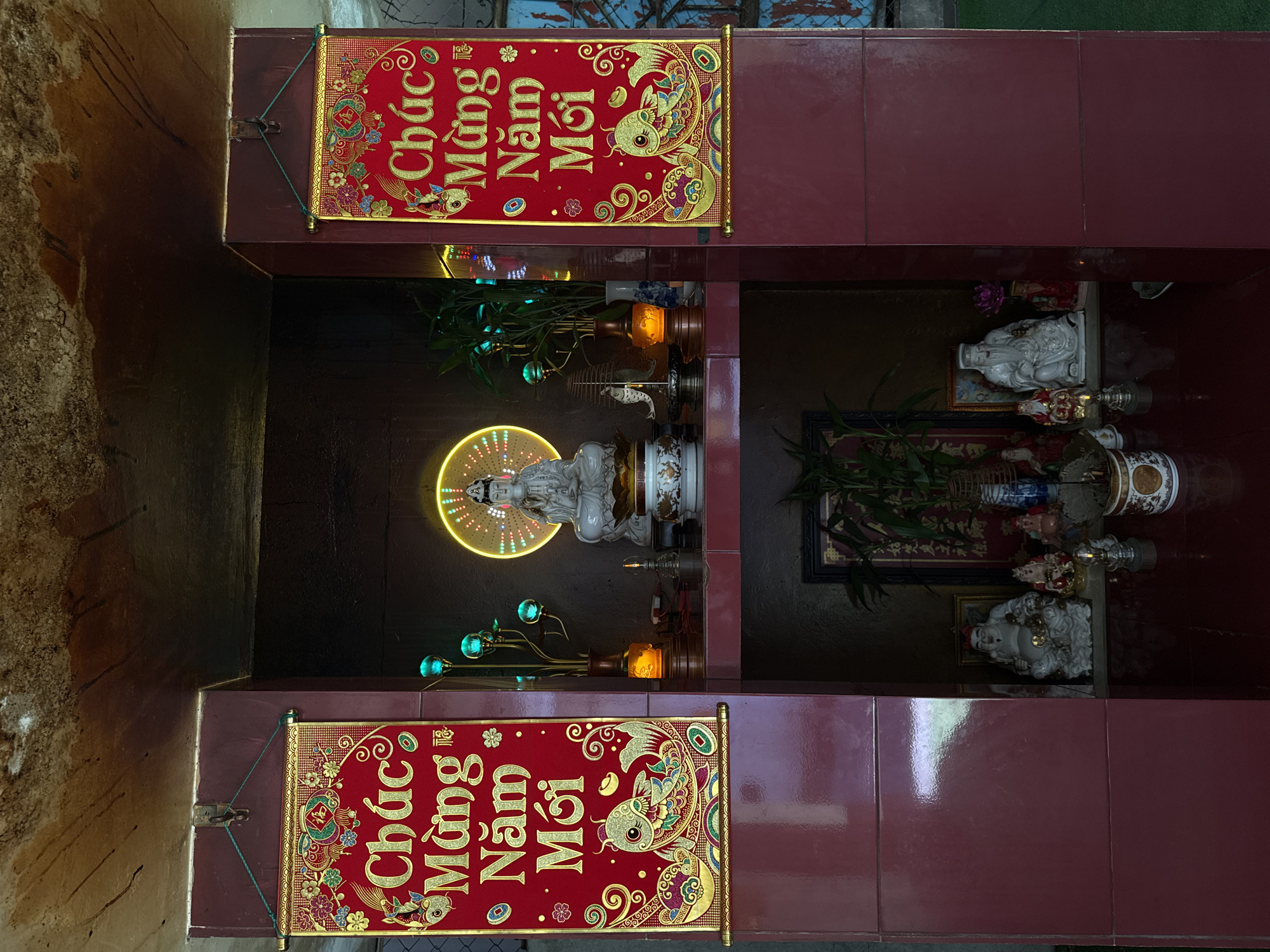 A small Buddhist shrine is set into a wall in the city of Nha Trang Ward, Vietnam. The shrine is red and brown. Two hanging banners, printed with Vietnamese text meaning "Happy New Year," frame a central niche. Inside the niche, a haloed statue of Buddha sits on a pedestal. A lamp resembling a bouquet with blue bulb-like flowers flanks the Buddha. Other statues are visible in the shadows.