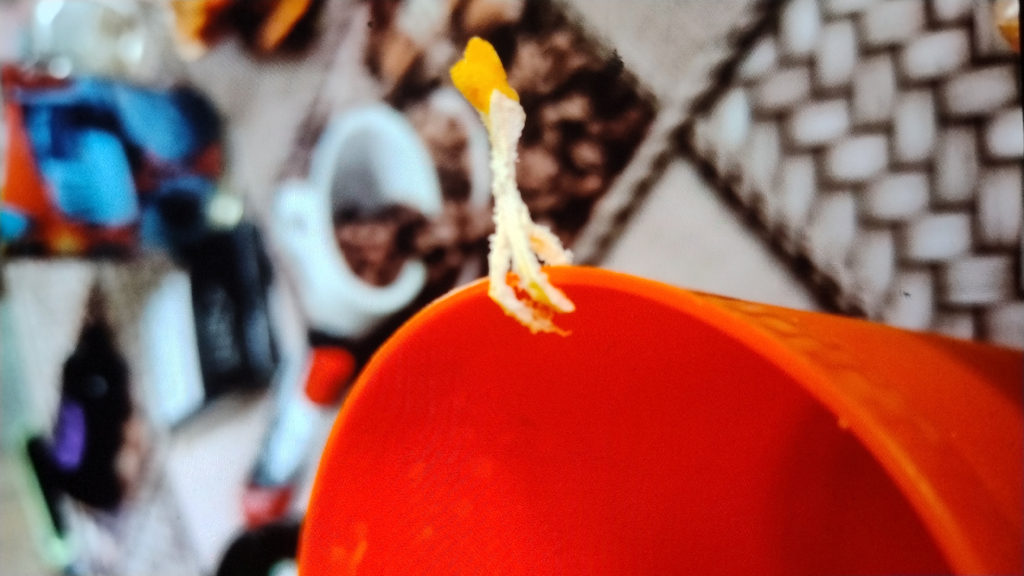 This is a close-up, low-resolution photograph of a portion of a table with various objects. In the foreground, a bright orange cup is visible on the left side of the frame. Emerging from the rim of the cup is a small, delicate, and somewhat wilted flower or plant stem with white petals, and a hint of yellow and red within its structure. The background of the image shows a patterned surface that appears to be a tablecloth or placemat with a woven texture in shades of beige and brown. Scattered on this surface are several out-of-focus objects, including what looks like a black and white rectangular package, and a colorful container or bottle with abstract designs in blue and orange. The overall impression is of a casual, indoor setting, possibly a home or a casual dining area. There is no information in the image to indicate a specific location in Mykolaiv, Ukraine.