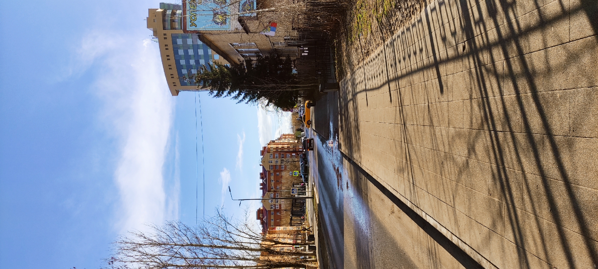 The image depicts a street scene in Fryazino, Russia. The camera is positioned on the edge of a sidewalk, which is constructed of rectangular paving stones. Shadows from a metal fence or railing are cast across the sidewalk. A row of buildings lines the opposite side of the street, which is partially wet, reflecting the sky. Cars and a yellow taxi are visible on the street. The sky is light blue with streaks of white clouds. Leafless trees are present along the sidewalk. A tall building with a partially visible sign reading "УСПЕХ" (Success) is on the left.