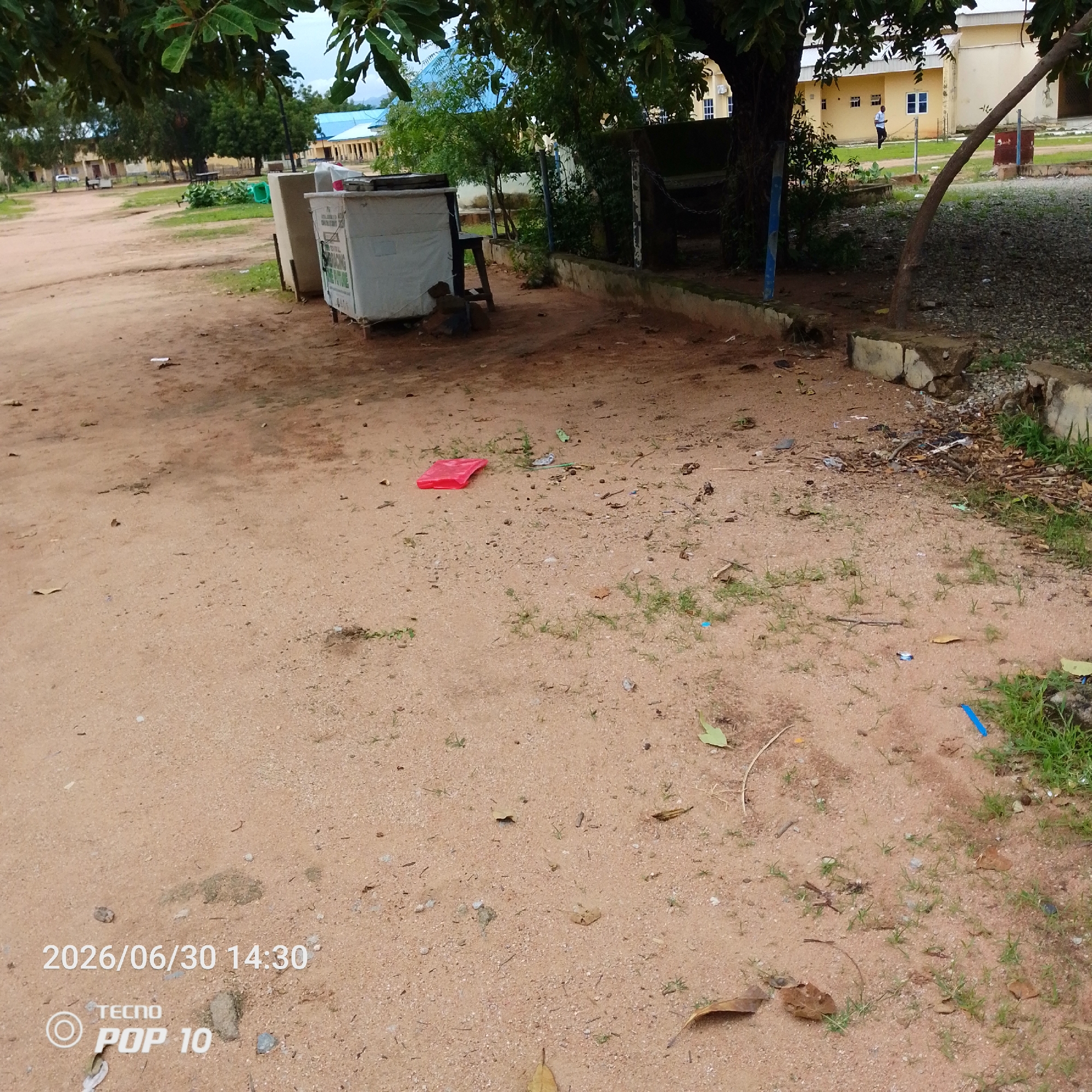 This media file shows an outdoor scene in Jalingo, Nigeria. The foreground is dominated by sandy ground with scattered debris, including leaves, twigs, and small pieces of trash. A bright red plastic bag lies prominently on the ground in the mid-left. In the mid-ground, there is a white kiosk or stall with some writing on it, and a dark stool leaning against it. Beyond the kiosk, there is a low concrete wall.
In the background, several buildings with yellow walls and blue roofs are visible. Trees with green foliage are scattered throughout the scene, casting shadows and contributing to a natural, somewhat unkempt environment. Some of the trees have large leaves and appear mature. A chain-link fence is partially visible. The ground cover in the background consists of gravel and patches of grass. The lighting suggests it is daytime, possibly late afternoon given the shadows. There is no clear indication of specific activity or emotion, presenting a static view of the location.
Visible text includes a timestamp at the bottom left of the image: "2026/06/30 14:30", and branding that reads "TECNO POP 10".