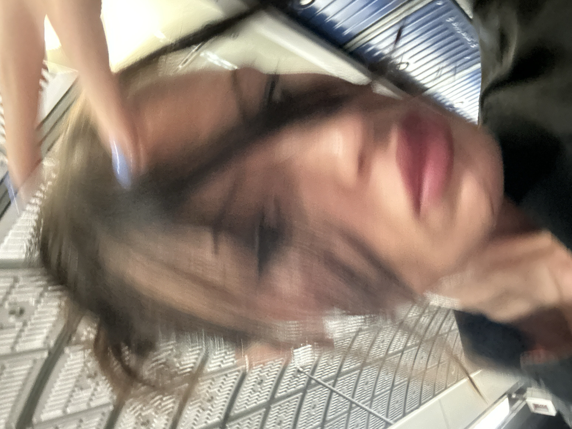 The image shows a blurred close-up of a person's face. The individual has closed eyes, visible lips, and dark hair obscuring part of their face. The background is a blurry mix of gray and white textures, with hints of blue in the upper portion. There is a suggestion of the person being inside what appears to be a shopping cart. The location is potentially inside a grocery or retail establishment. Due to the blurry nature of the image, specific details about the person's identity, the exact location in the UK, or the exact nature of their interaction with their environment are unclear.