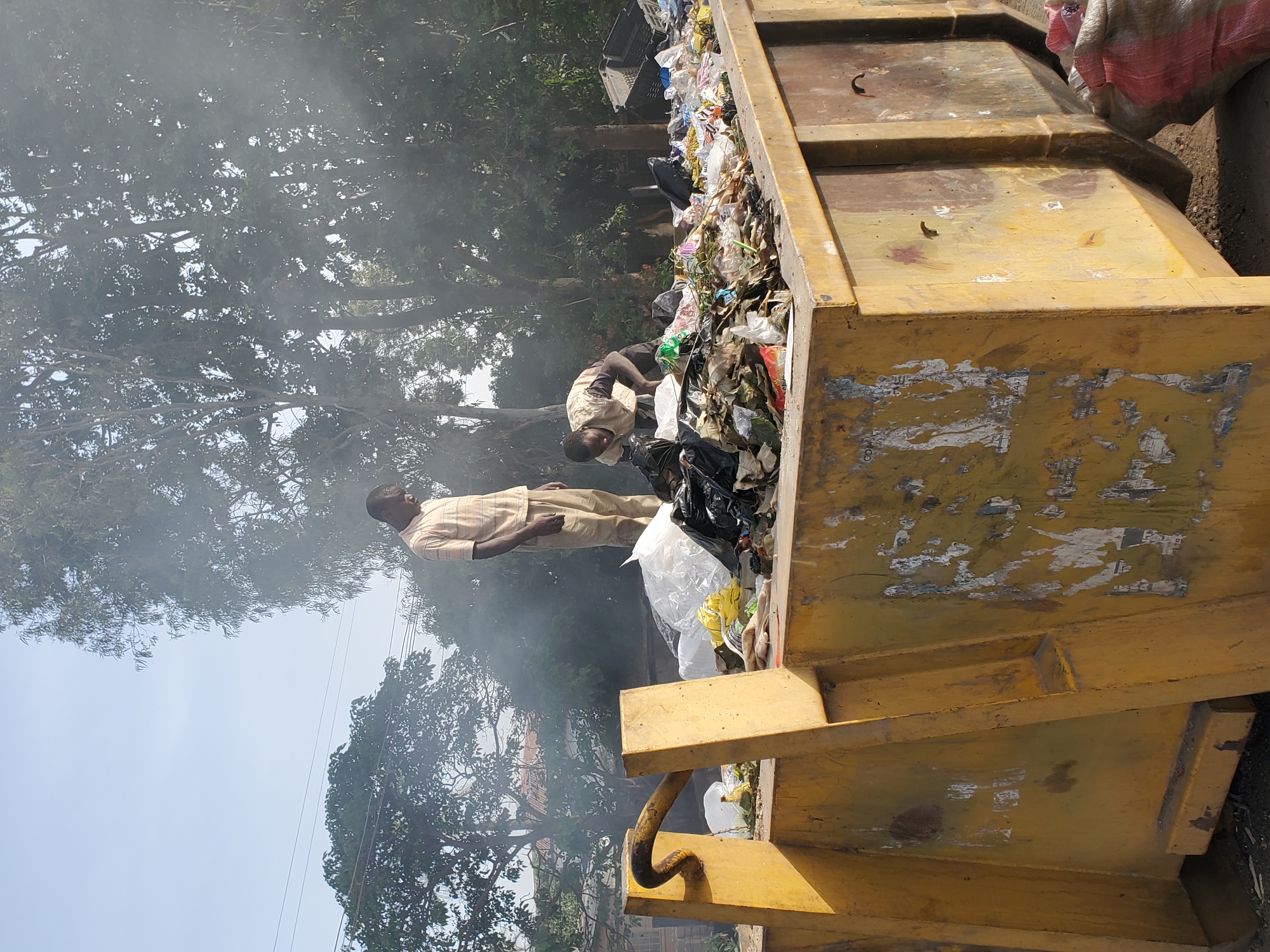 The image captures an outdoor scene in an urban or peri-urban setting, likely in Jos, Nigeria, centered around a large, yellow industrial dumpster overflowing with trash. Two men are visible interacting with the refuse. One man, dressed in a light-colored, possibly striped shirt and light trousers, stands upright to the left of the dumpster. Another man, wearing a beige or light brown shirt, is bending over the edge of the dumpster, appearing to be sorting through the contents or adding items to the already full bin.
The dumpster is packed with a diverse assortment of waste, including numerous plastic bags, wrappers, and general refuse, much of which spills out. The yellow paint of the bin is visibly worn and scuffed, with remnants of torn stickers or labels, though no legible text is discernible.
A prominent feature of the scene is the significant amount of smoke or haze that permeates the air, particularly in the background, partially obscuring the lush green foliage of large trees. This smoke appears to emanate from the general area of the dumpster and surrounding vegetation, giving the scene a diffused and hazy atmosphere. The sky, visible in parts through the trees, indicates it is daytime. The men's expressions are not visible, but they appear focused on their task near the dumpster.