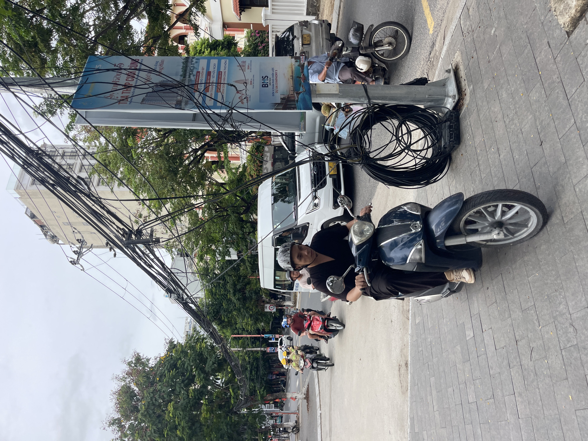 A street scene in Da Nang, Vietnam, features a man on a dark blue scooter in the foreground, wearing a black shirt and helmet. Behind him, several other scooters and a white van are visible on the road, with people riding them. A tall utility pole with a large bundle of black wires and a blue advertisement poster is located on the right side of the frame. The sidewalk is paved with gray rectangular tiles. Trees and buildings line the street in the background, under an overcast sky. The advertisement poster appears to be for a residential development called "Symphony 5" and includes Vietnamese text and images of buildings and water.