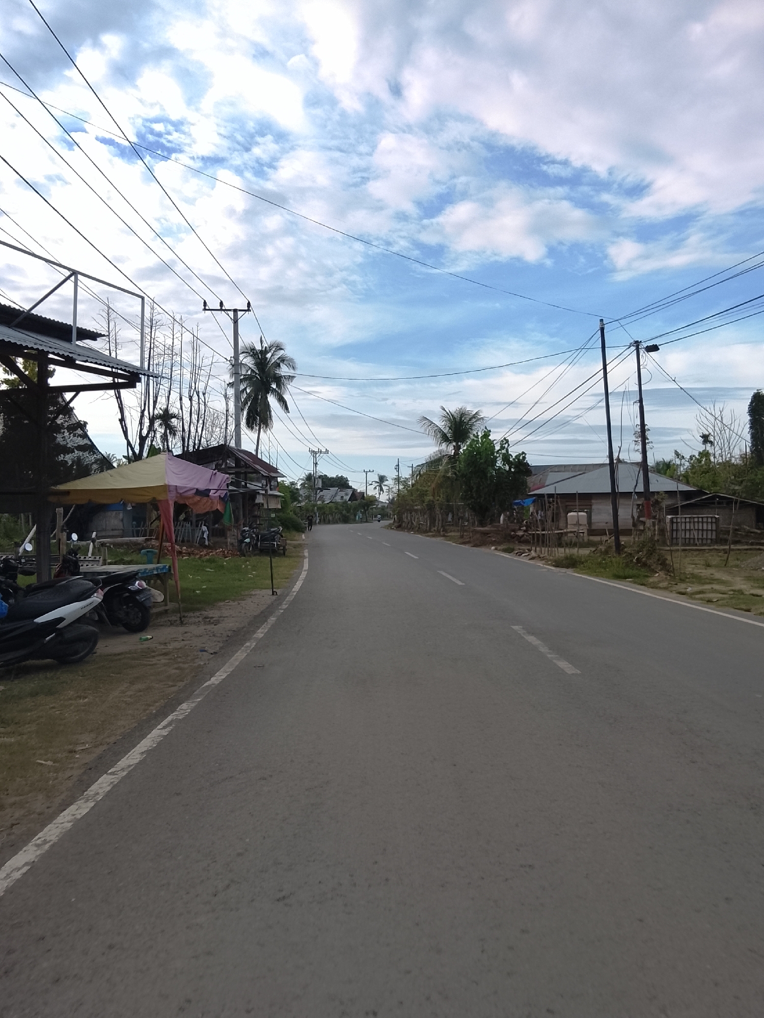 The image depicts a street scene in Blang Cut, Indonesia, under a partly cloudy sky. The road, asphalted and marked with a white dashed line, stretches into the distance, bordered by vegetation and scattered buildings on either side. On the left side of the road, a small market stall with a yellow and pink canopy is set up, displaying some laundry. Several motorcycles are parked in front of the stall. Beyond the stall, there are more structures, some appearing to be simple houses with wooden walls and corrugated metal roofs, interspersed with trees, including palm trees.
On the right side of the road, more houses and some vegetation are visible, with utility poles carrying numerous wires dotting the landscape. The overall impression is of a rural or semi-rural residential area. The lighting suggests it is either early morning or late afternoon, with the sun not at its highest point, as evidenced by the shadows and the general brightness. The presence of utility poles and the style of the houses indicate a developed, albeit simple, infrastructure. No specific activity or event is clearly depicted, other than the general presence of a road with passing traffic (implied by the road itself and a distant figure or vehicle on the right) and parked vehicles. The scene is peaceful and functional, reflecting everyday life in this Indonesian location.