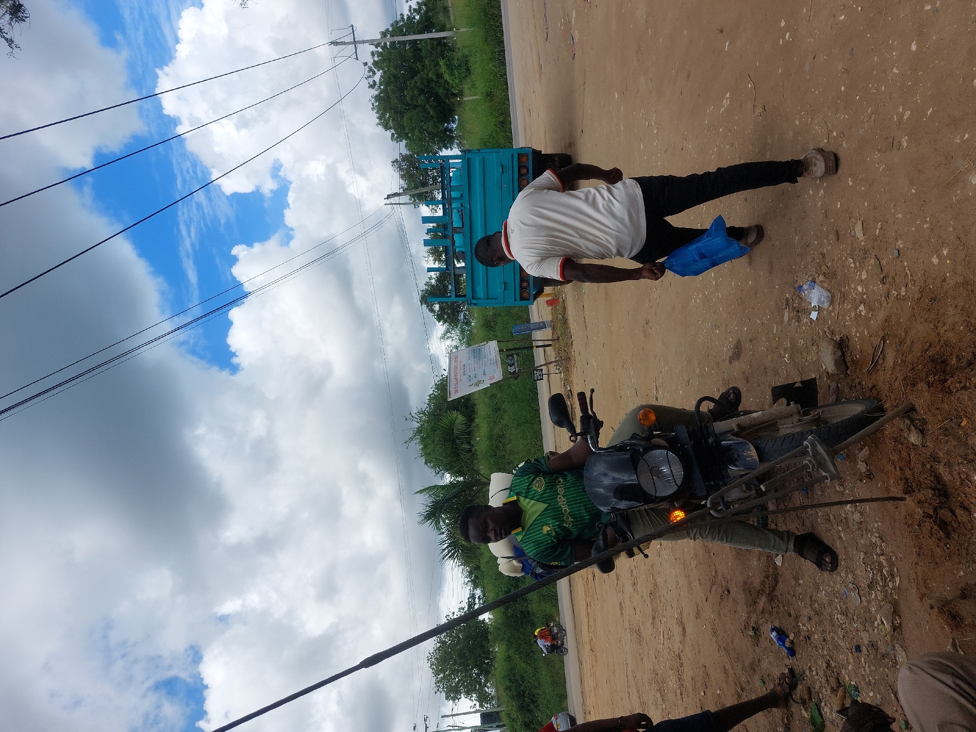 The image shows a street scene in Dar es-Salaam, Tanzania. A man in a green and yellow jersey sits on a motorcycle, with various metallic farm tools attached. Another man, wearing a white shirt and black pants, walks nearby carrying a blue plastic bag. A light blue truck is parked in the background, near a sign with text written in Kiswahili. The ground is sandy and littered with debris. Power lines stretch across the top of the frame, and the sky is partly cloudy.