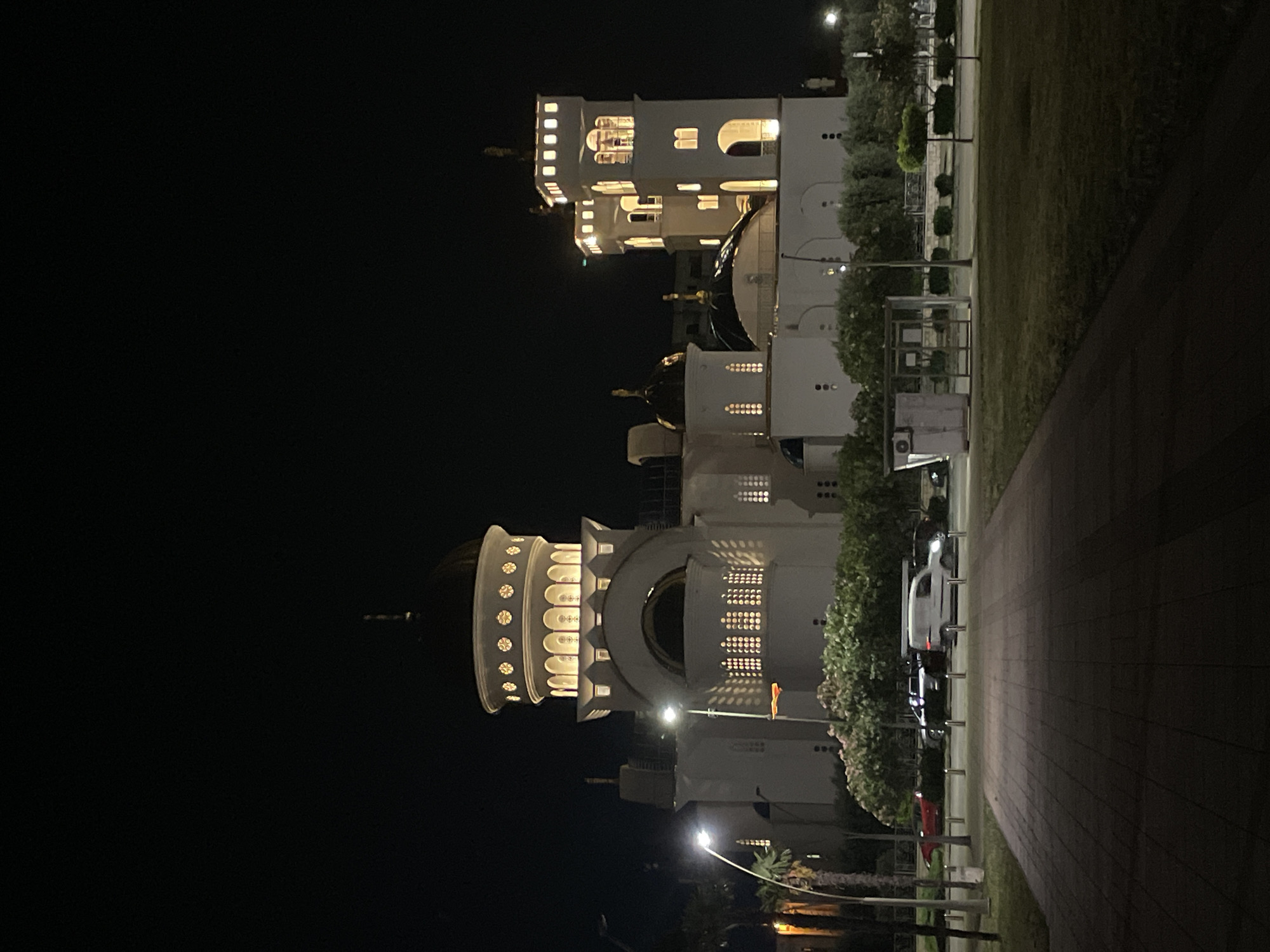 The image captures the illuminated dome and facade of a building at night, likely a religious or governmental structure due to its architectural style. The building's exterior is white, adorned with circular patterns and arched windows glowing with warm light. Adjacent to the building, a paved pathway runs along a grassy area. Cars are parked along the side of the path. The sky is dark, indicative of nighttime, with artificial lights scattered around, illuminating the scene. The image, given the prompt context, is likely a view of the King Nikola's Palace (Dvorac Kralja Nikole) in Bar, Montenegro.
