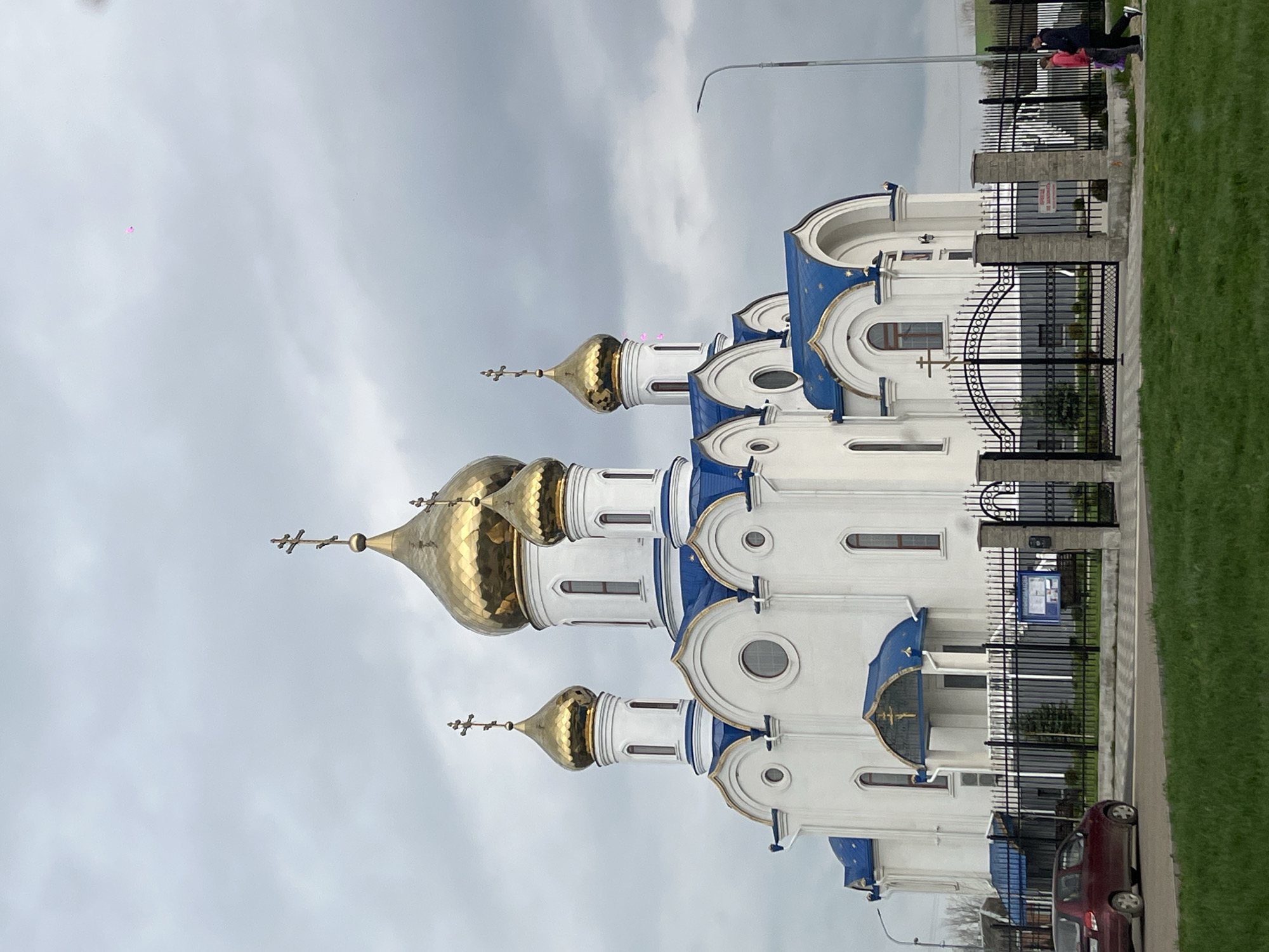The image captures a white and blue Orthodox church with golden domes, situated in what appears to be Novopokrovskaya, Russia. The church is enclosed by a black wrought-iron fence. Two individuals are visible walking in the background along the sidewalk. A parked red car is near the church. The sky is overcast, suggesting a cloudy day.