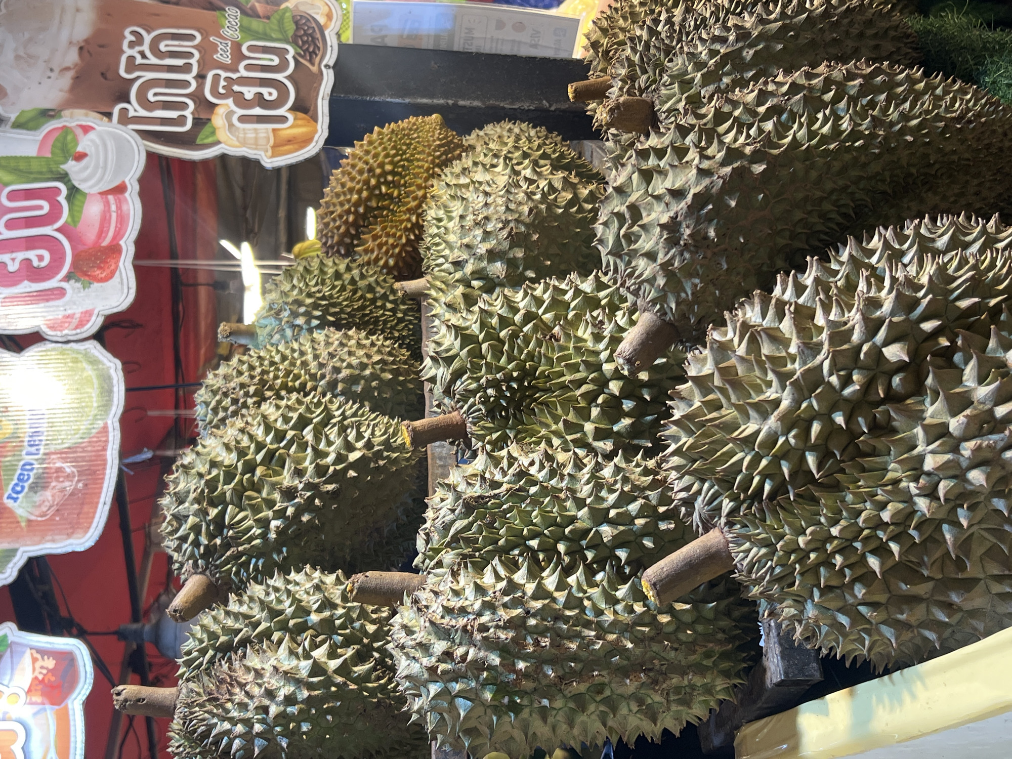 This is a close-up, eye-level, medium shot of a stall selling durian fruit in Karon, Thailand. The foreground and midground are filled with a dense arrangement of large, spiky durian fruits, predominantly green and brown in color. Several durians are piled on top of each other, with their characteristic thorny exteriors clearly visible. The stems of some fruits protrude upwards. In the background, partially visible above the fruit, are colorful signs featuring Thai text and imagery of drinks and desserts, suggesting a food stall or market setting. One sign prominently displays an image of a chocolate drink with whipped cream and the Thai words "โกโก้เย็น" which translates to "iced cacao." Another sign features images of macarons and strawberries. The lighting appears to be natural or from overhead market lights, casting some shadows and highlighting the textures of the fruit. The environment appears to be outdoors, typical of a street market or roadside stall. There are no people visible in the frame.