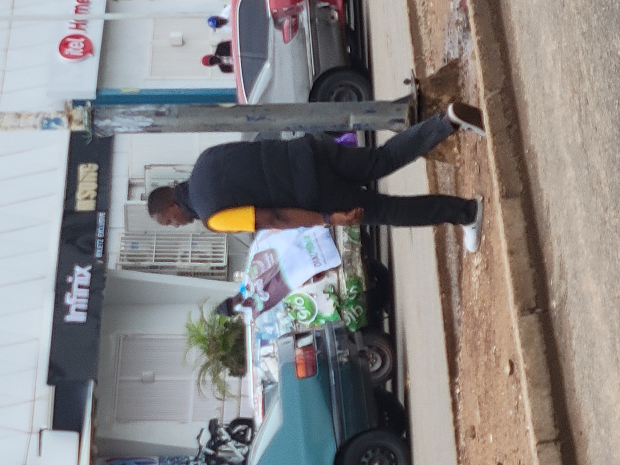 A man in a black shirt with a yellow armband, black pants, and white sneakers is walking on a sidewalk next to a street in Jos, Nigeria. He is holding a stack of papers with a "Dial 9mobile" logo and advertisements on top of a car. The car is parked near buildings that have signs reading "Infinix," "KONSUMS," and "itel Home." A variety of vehicles, including cars and motorcycles, are parked in the vicinity. The weather appears sunny.