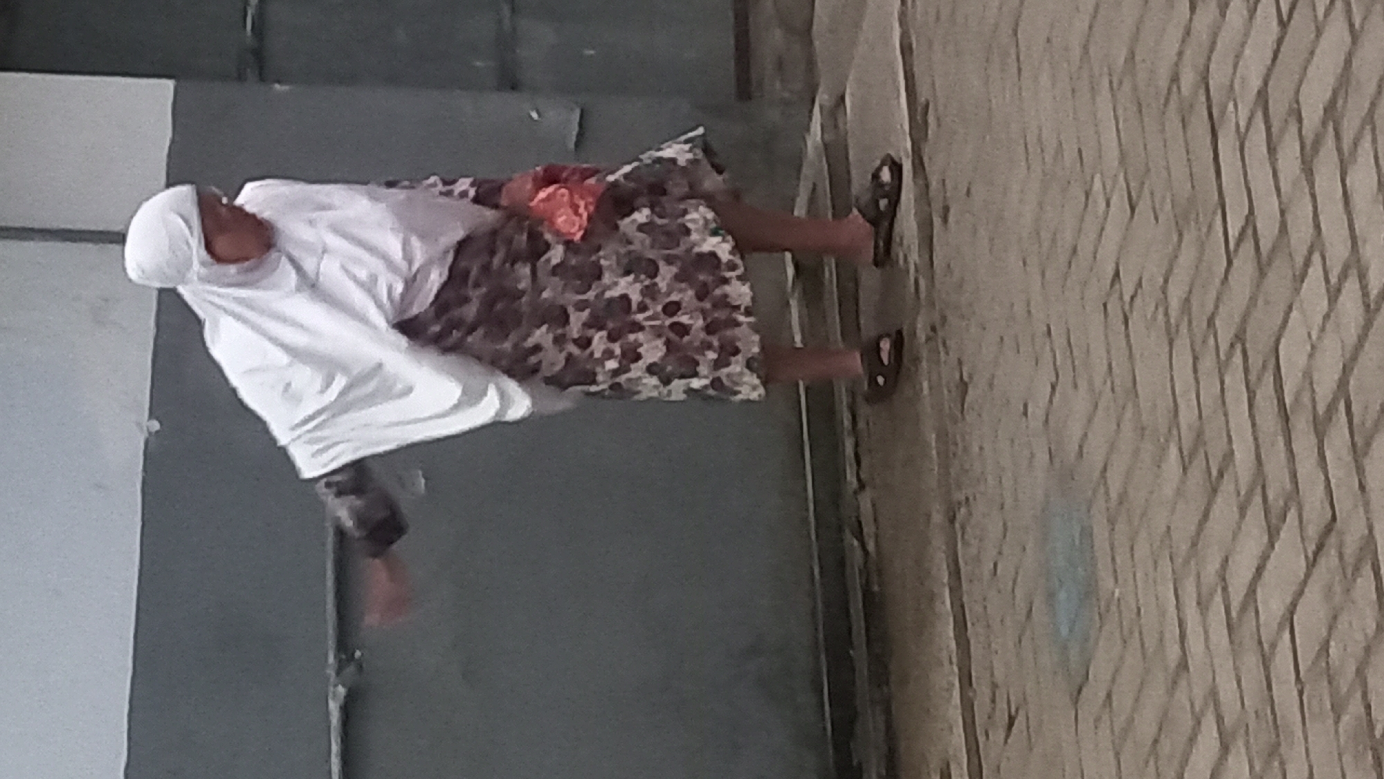 An elderly woman wearing a white hijab, a patterned dress, and black sandals is standing on a brick-paved walkway in front of a gray wall. She is holding a pink cloth object in her right hand and her left arm is extended. The scene is an outdoor environment in the city of Pare, Indonesia.