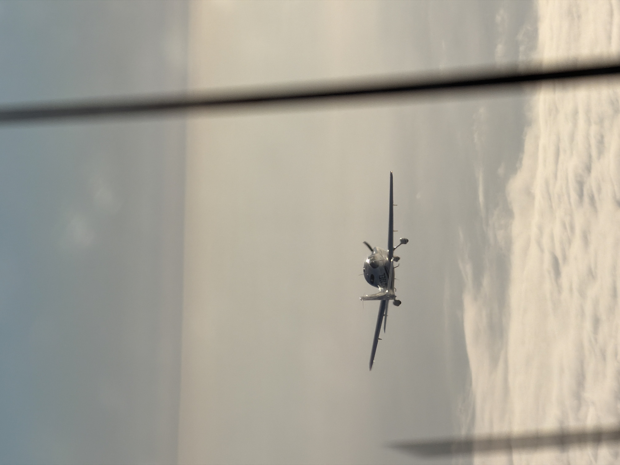 An aerial view shows a small white airplane flying above a thick layer of clouds. The plane is viewed from a slightly below and to the side angle. The sky above the clouds is a pale blue with some hazy cloud formations. The plane has the registration number "N219EL" visible on its tail. The image also shows a blurry, vertical dark object on the right side of the frame, possibly a part of a structure or window frame. The scene is shot with a shallow depth of field, with the plane and the clouds in focus and the foreground blurry. This image was taken from the city of Ventura, United States of America.